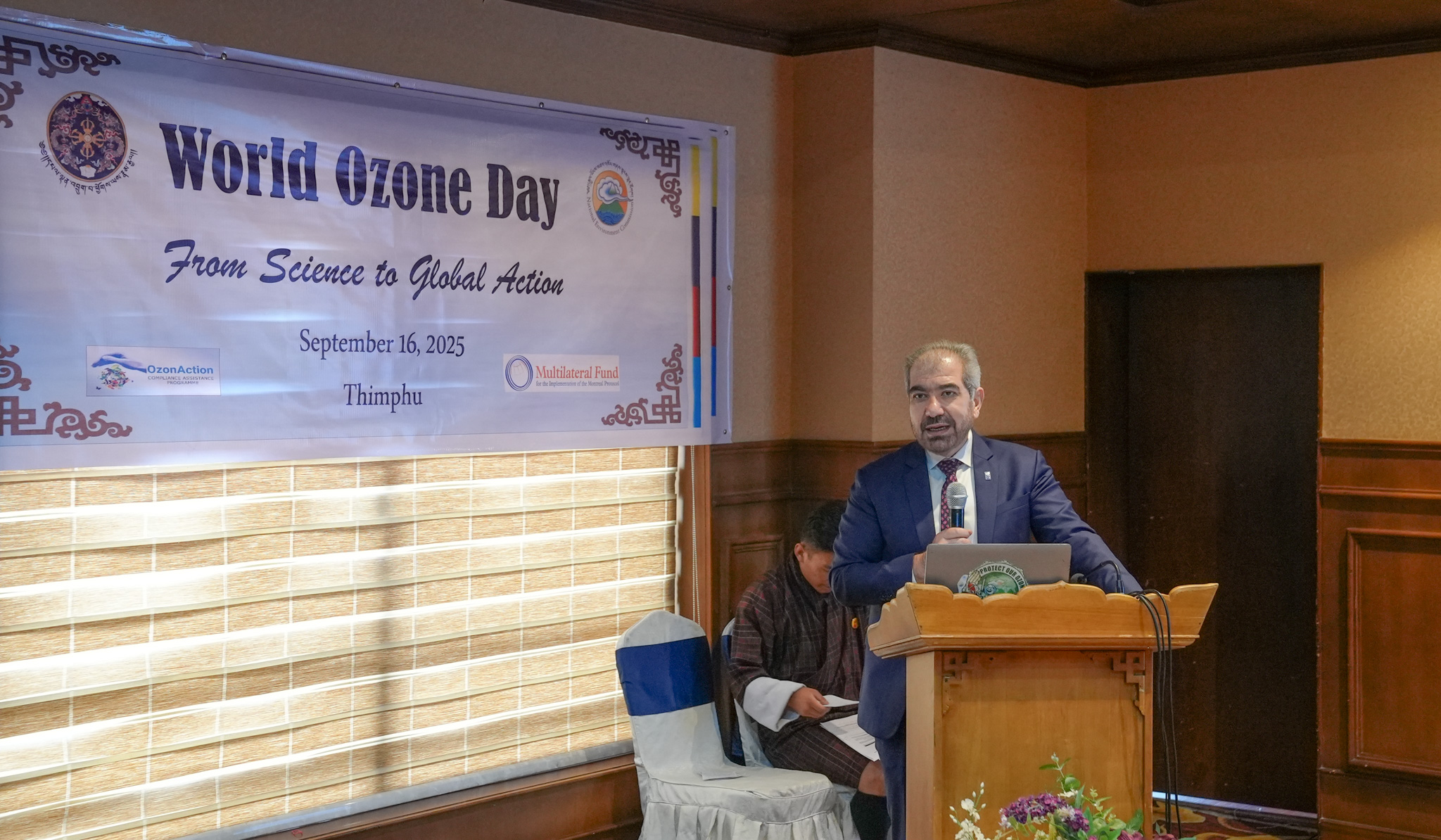 Photograph from World Ozone Day: man at the podium delivering speech, another person seated beside.