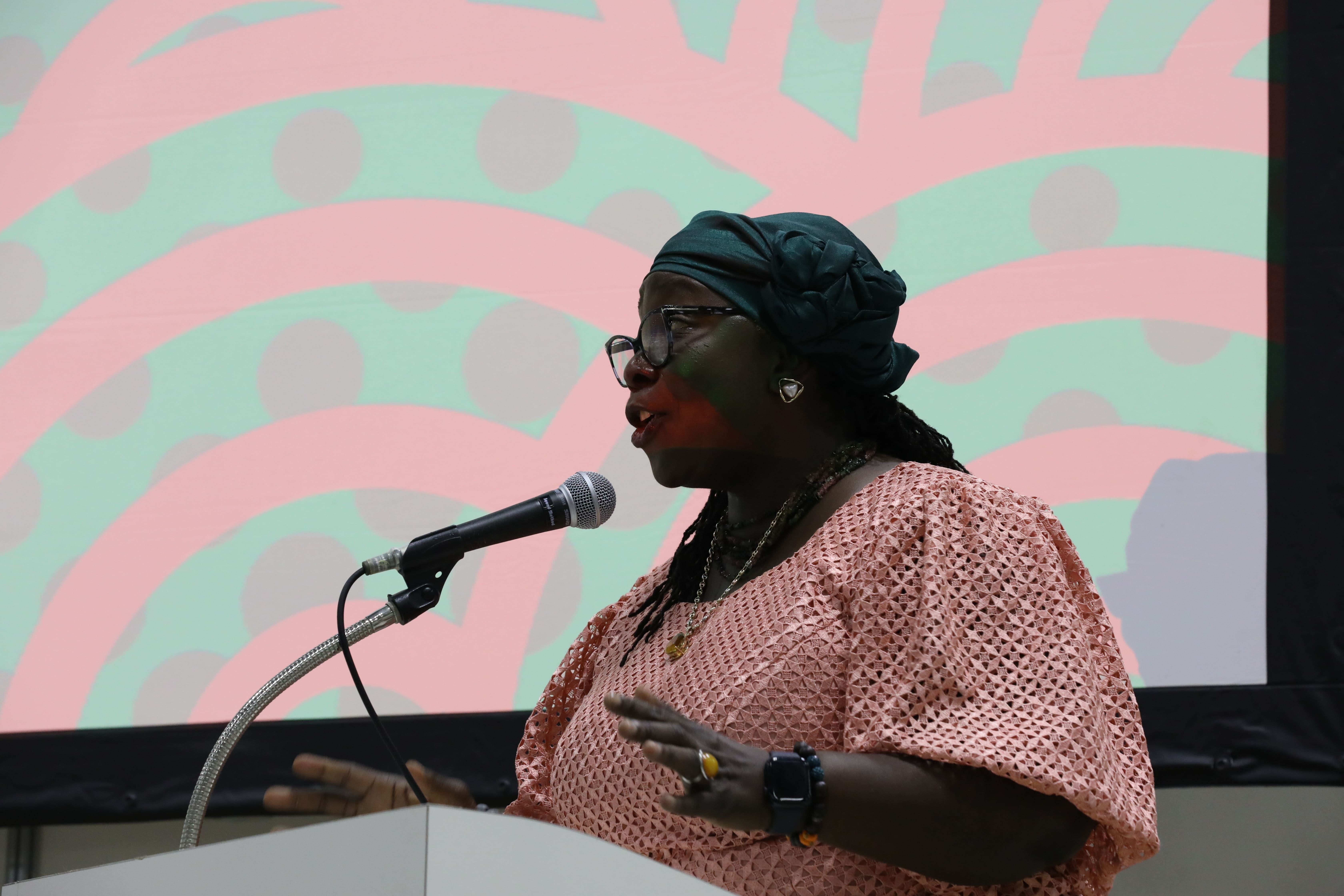 Speaker at podium with microphone, wearing a pink patterned top, colorful circular backdrop.