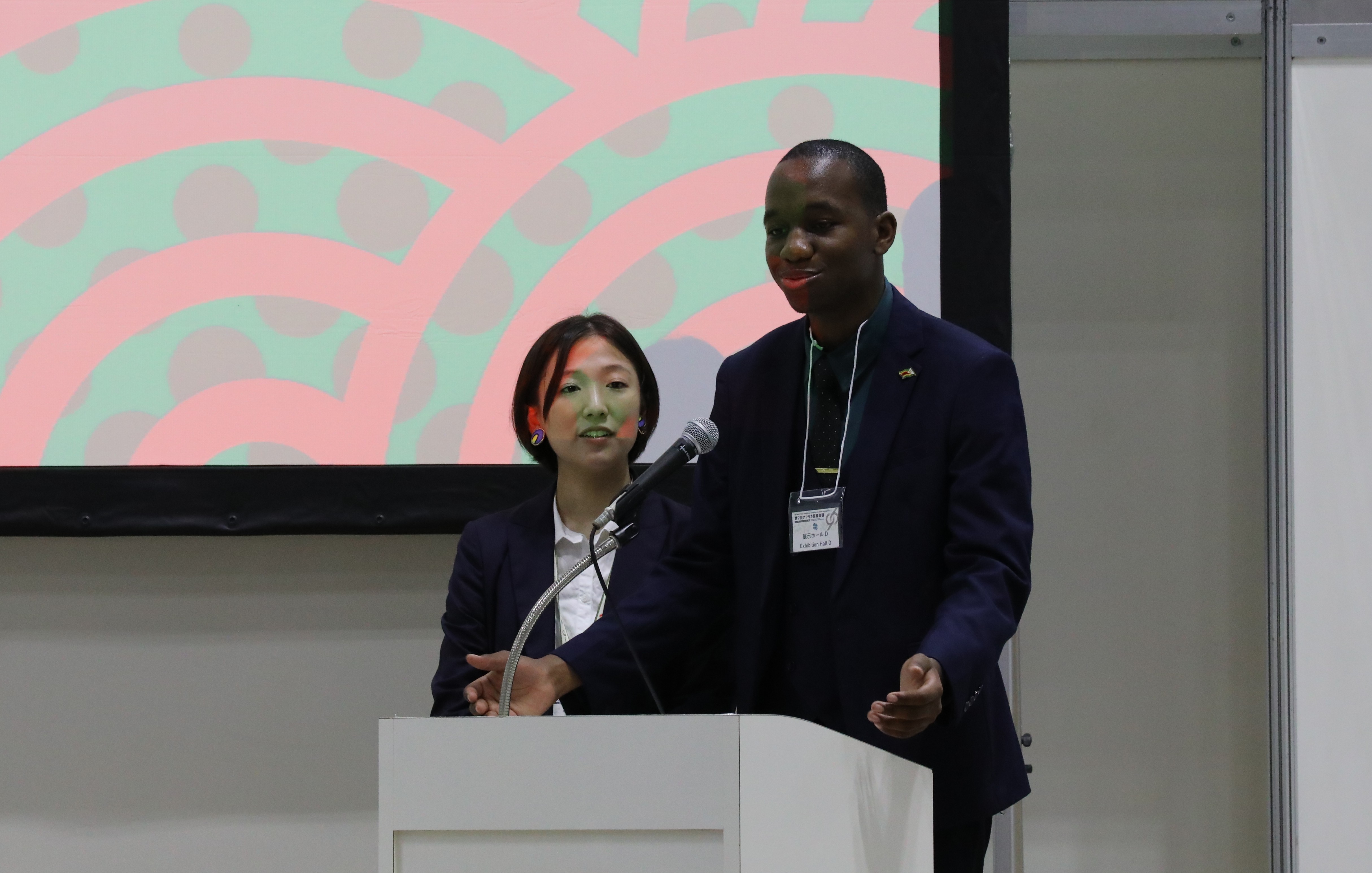 Two presenters stand at a white podium as a pink circular-pattern slide fills the screen.