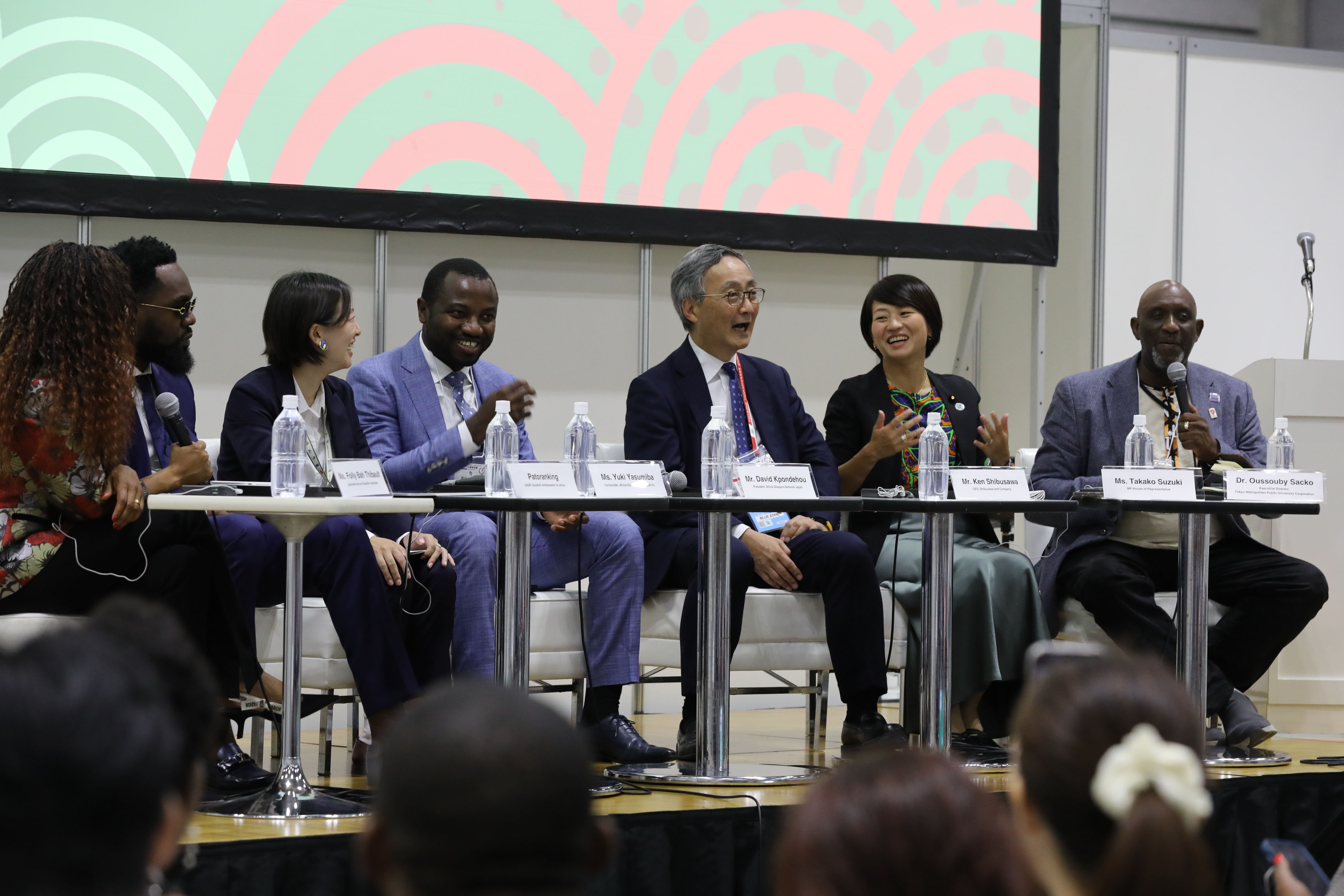 Photograph of diverse panelists seated at a long conference table on stage, audience in foreground.
