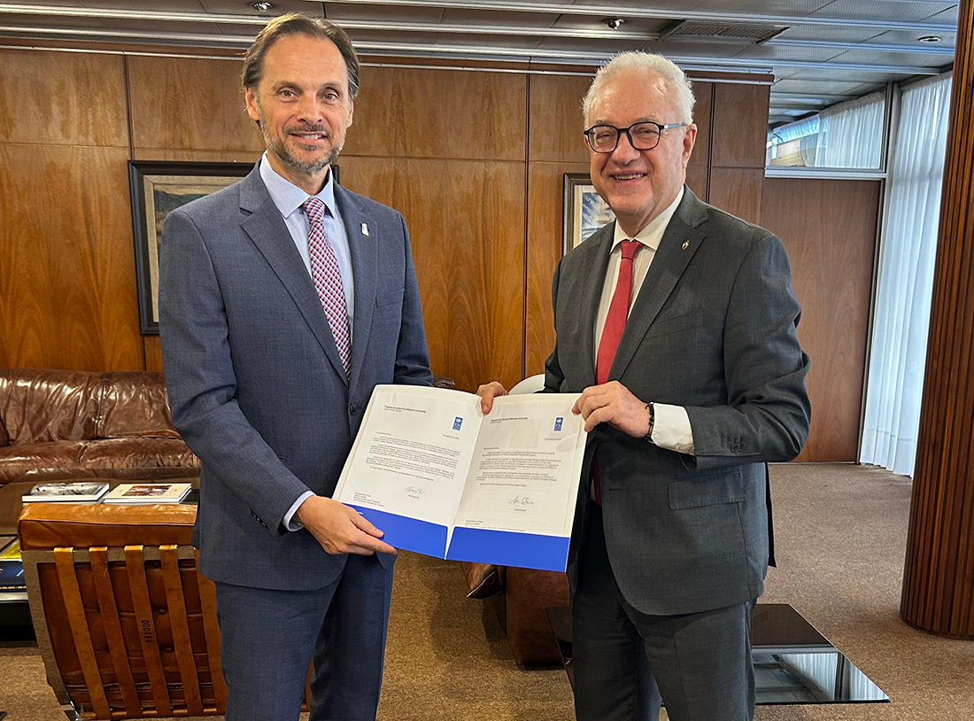 Two men in formal attire are smiling and holding documents in an office setting.
