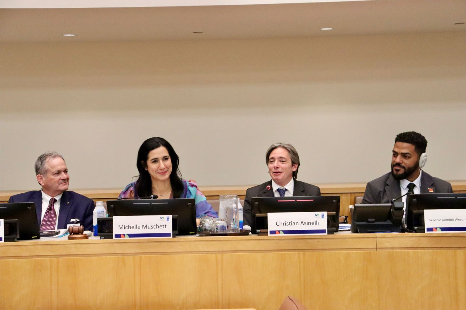 Five panelists sit at a long conference table with nameplates in front.