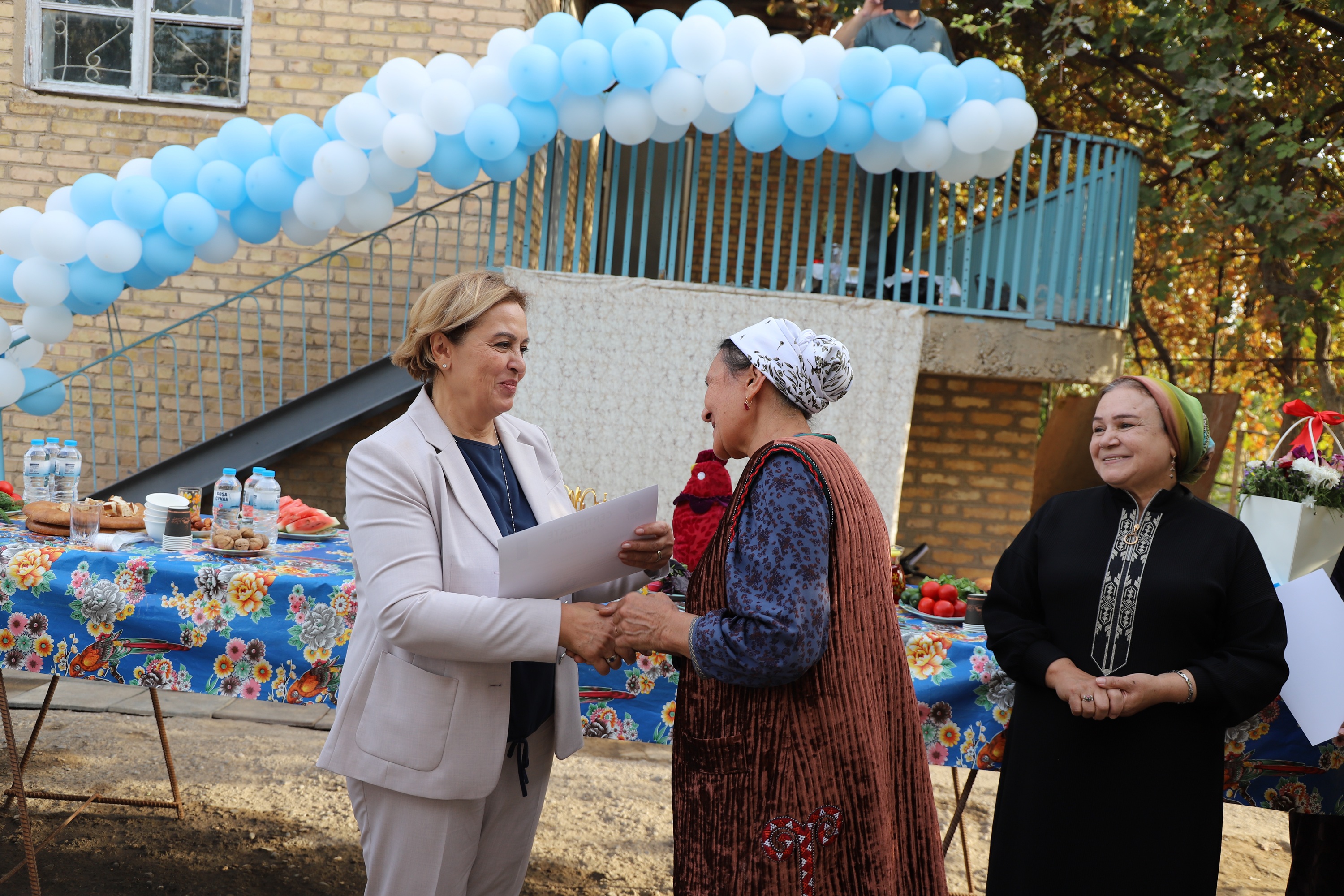 Three women under a blue-and-white balloon arch; a beige-suited woman greets an elderly woman.