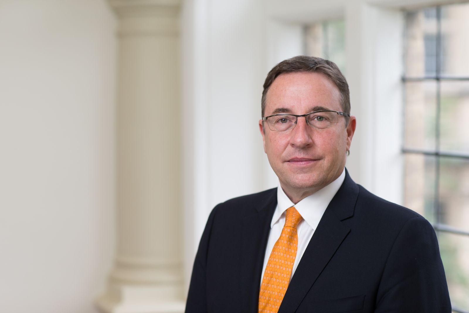 Man in a dark suit and orange tie stands indoors by a window, with columns in the background.