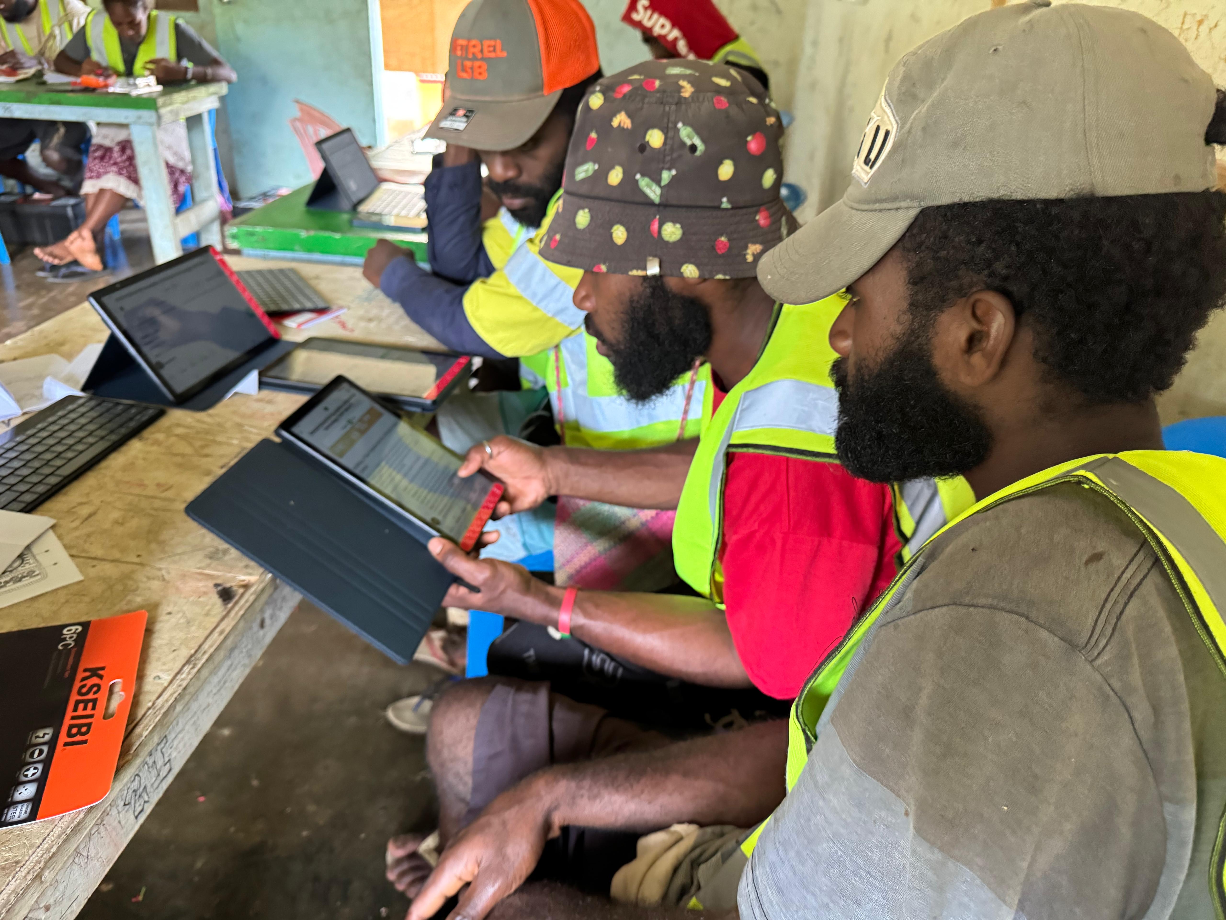 Photo: Group of people in hi-vis vests and hard hats using laptops at a workshop.