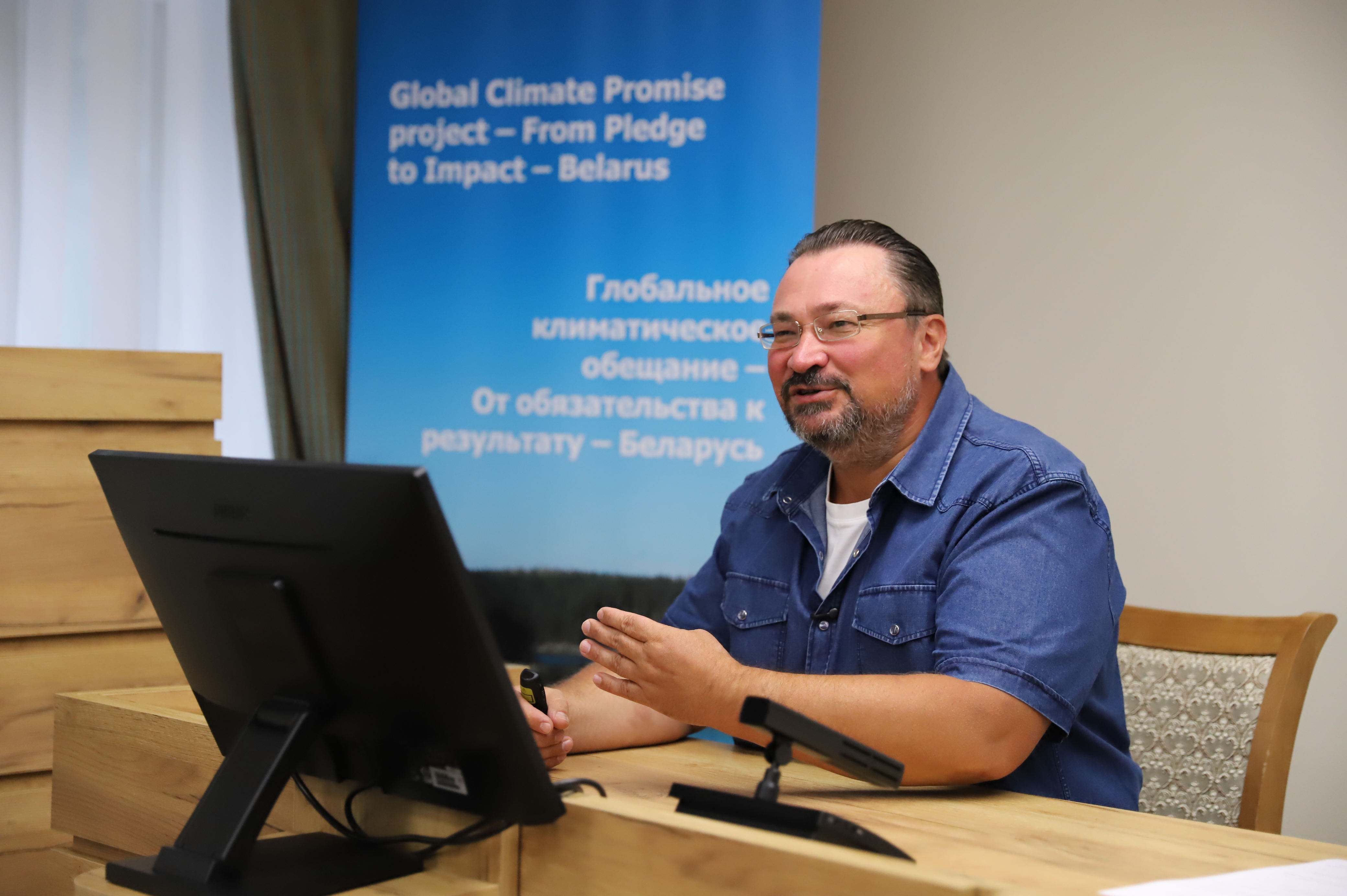 Person in blue shirt sits at desk with computer and microphone; blue banner in background.