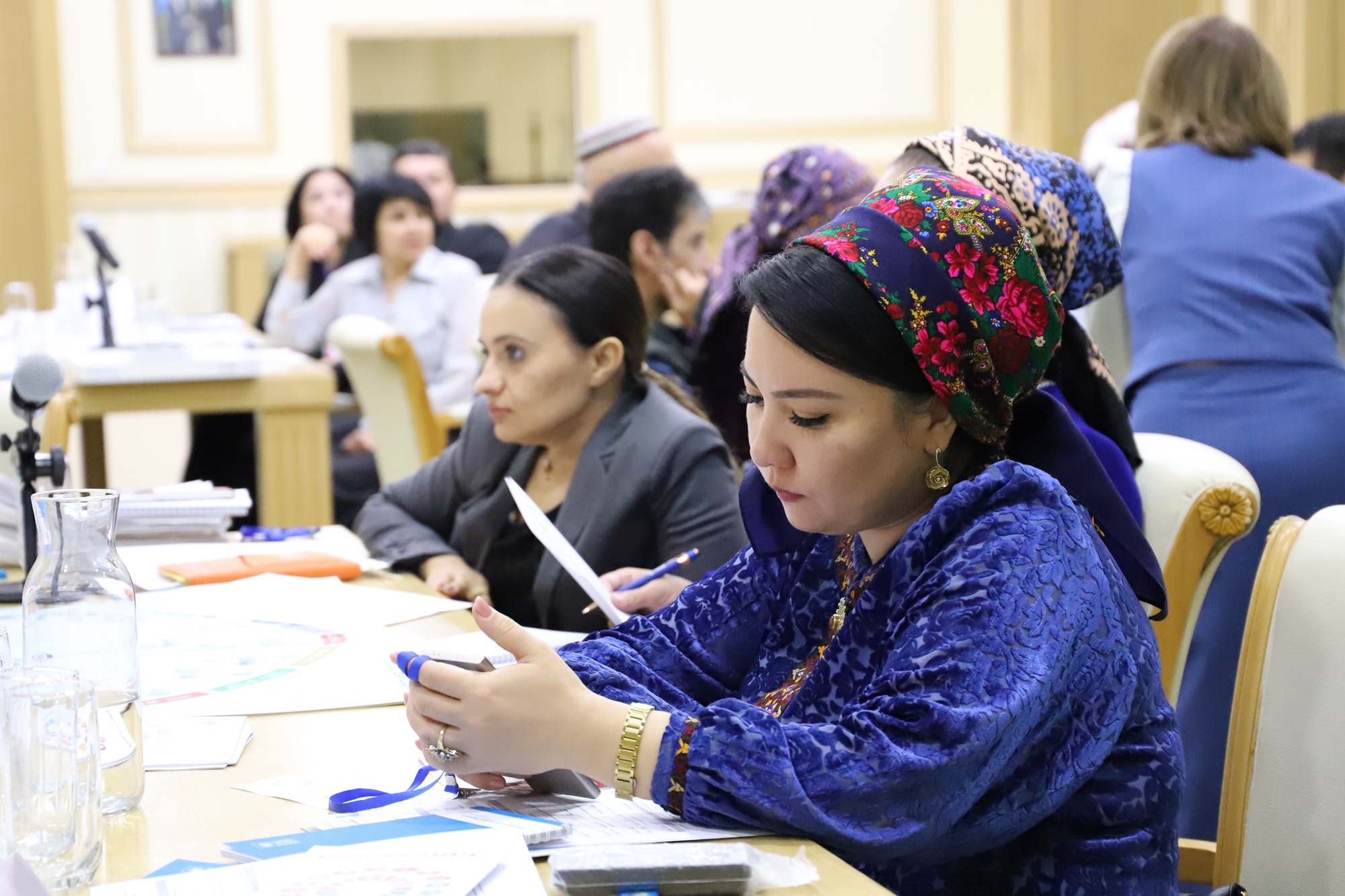 Photograph: workshop participants at long table; foreground woman in blue headscarf holding phone.