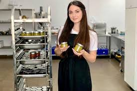 Cook in white shirt and dark apron stands in a kitchen with metal shelves, holding a yellow item.