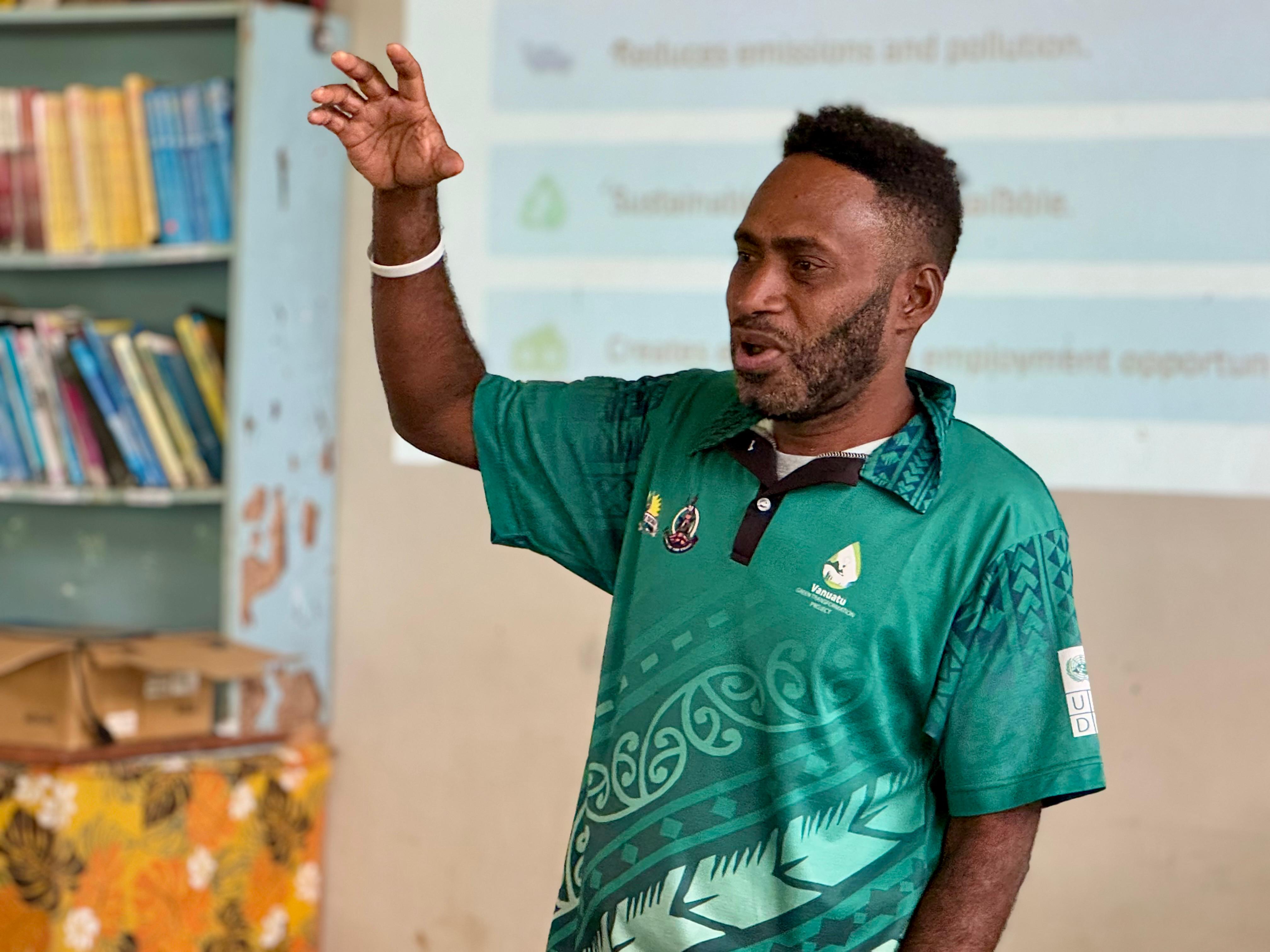Person raises hand in classroom; green patterned shirt, bookshelves and projector in back.