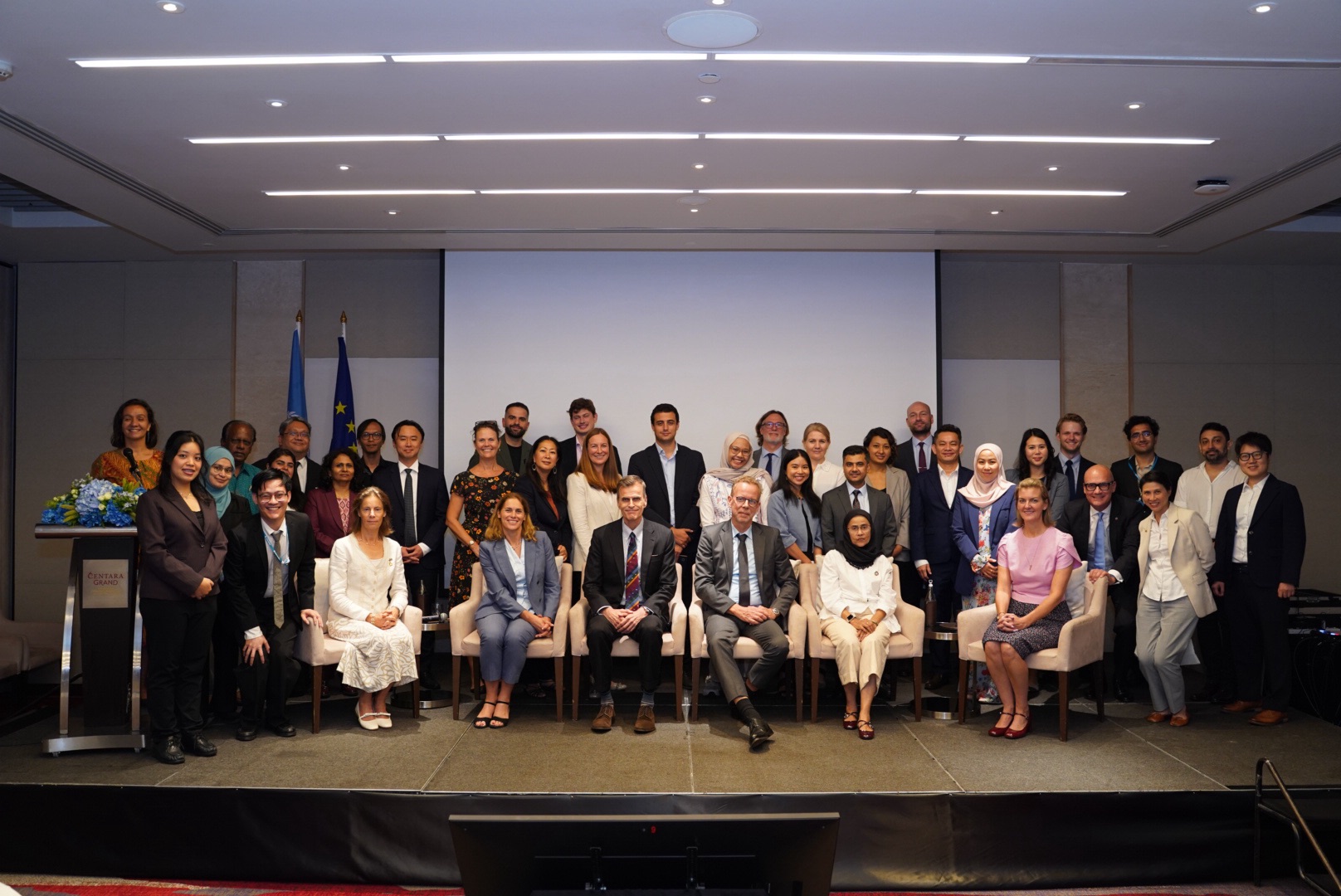 Group of diverse professionals posing on a conference stage with flags and screen.
