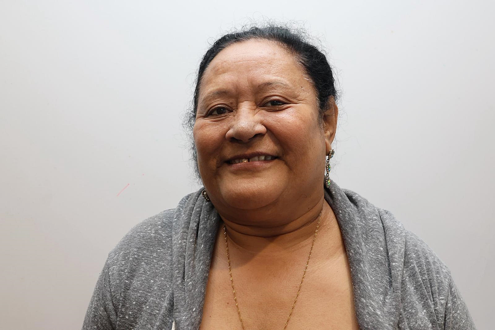 Person in gray cardigan over brown top with pendant necklace, standing against a light gray wall.