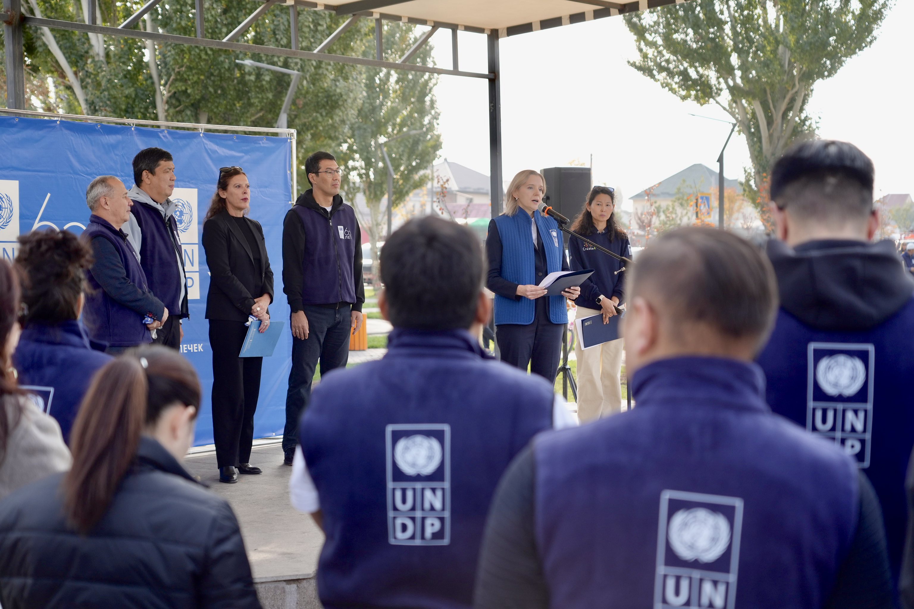 Outdoor UNDP event with panelists on stage and attendees in blue UN jackets.