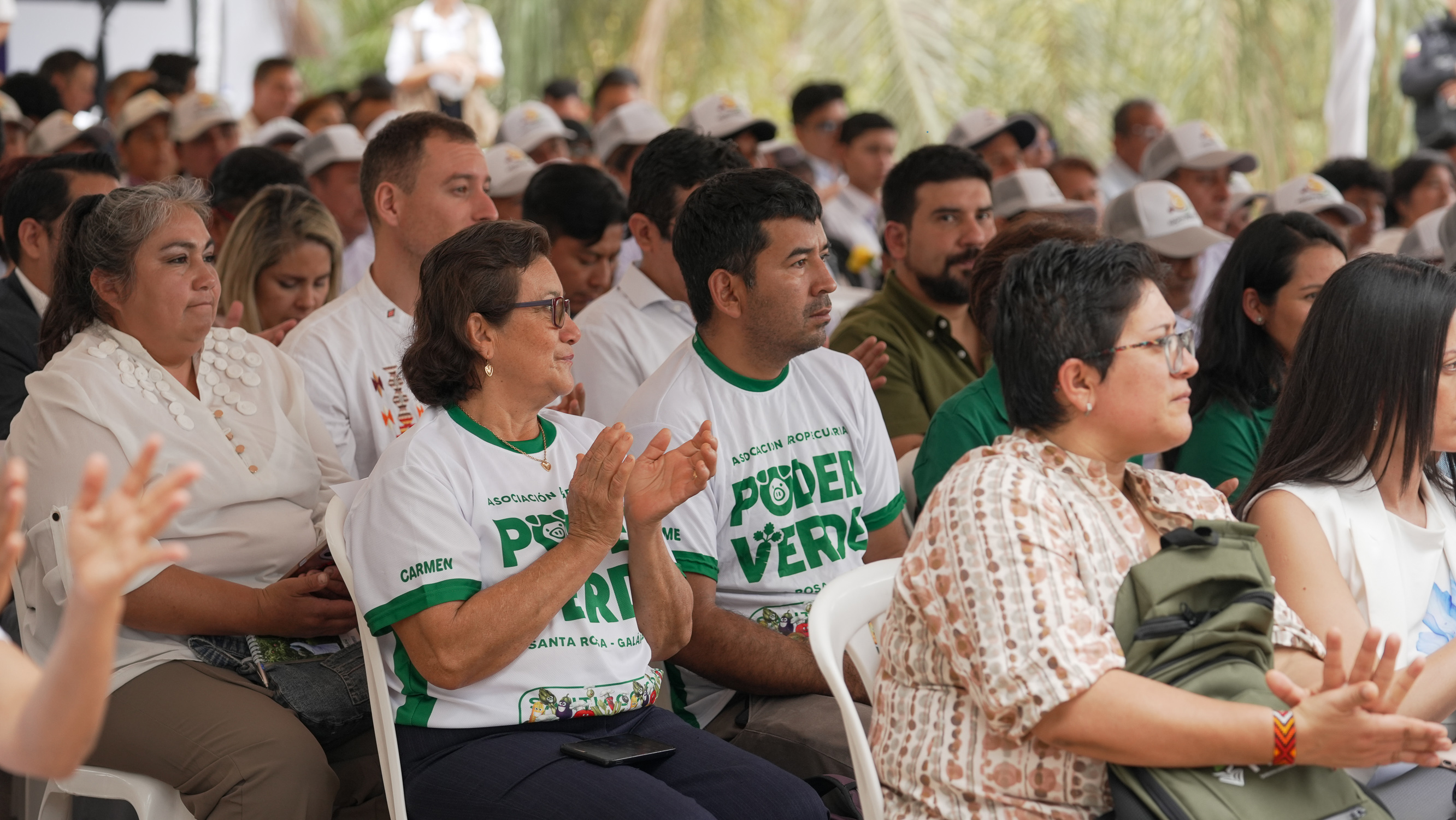 Outdoor crowd seated in rows, many wearing white shirts with green text, attentive.