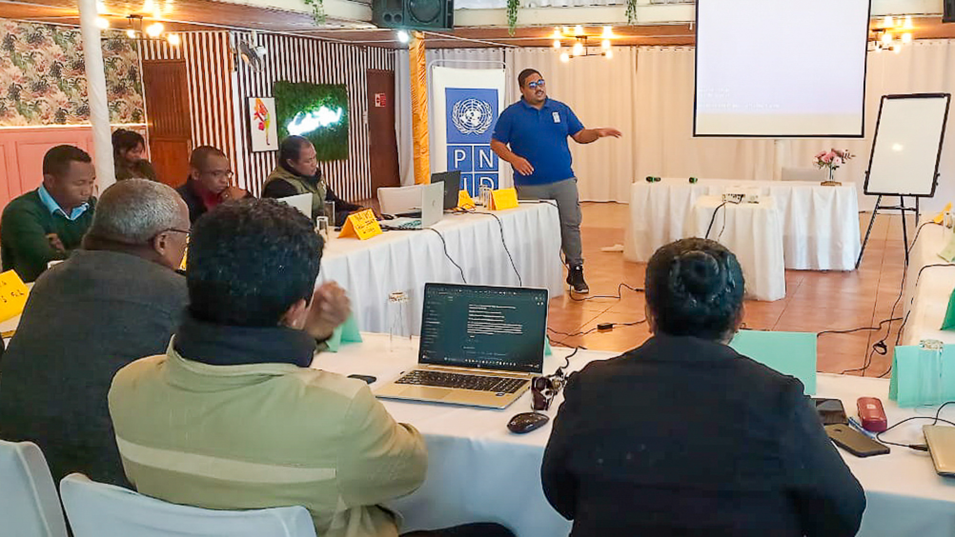 Presenter in a blue shirt speaks at the front of a conference room to seated attendees.