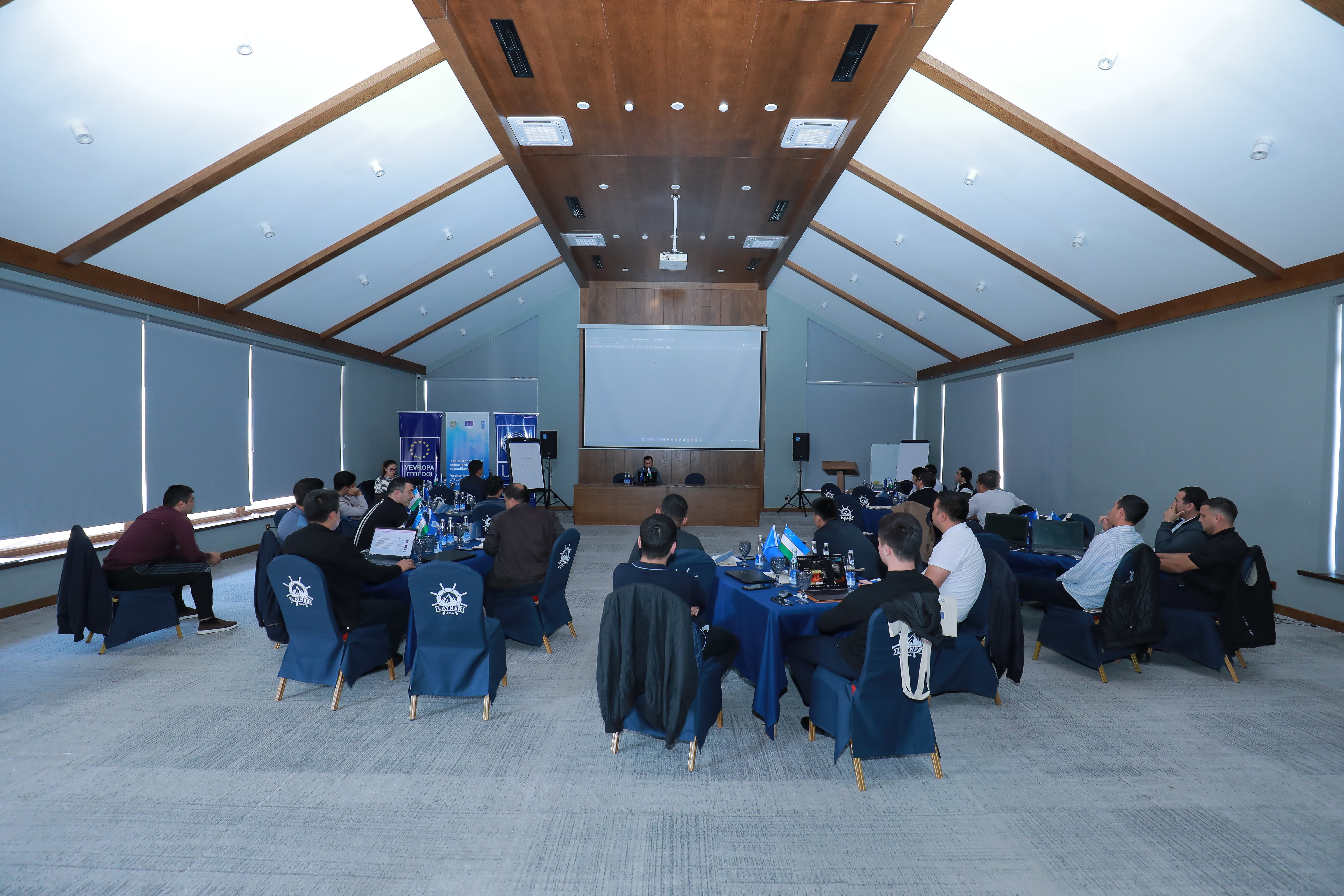 Photograph of a conference room with round tables, blue chair covers, and a large front screen.