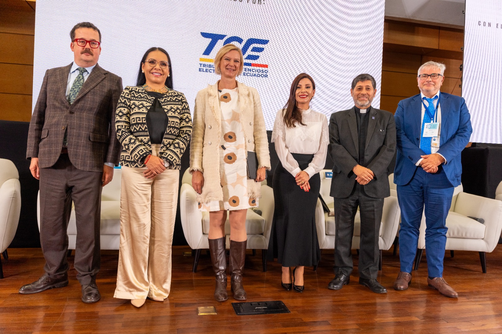Group of six professionals on stage at a conference, smiling, with a blue logo banner behind.