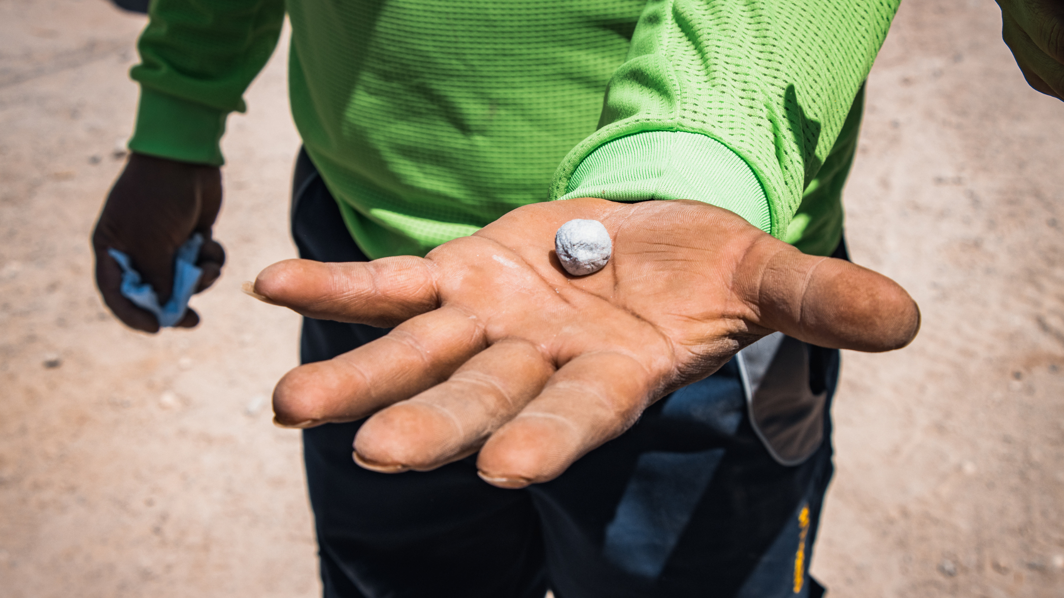 Person in green shirt extends hand toward camera, palm showing a coin; blue item in the other hand.