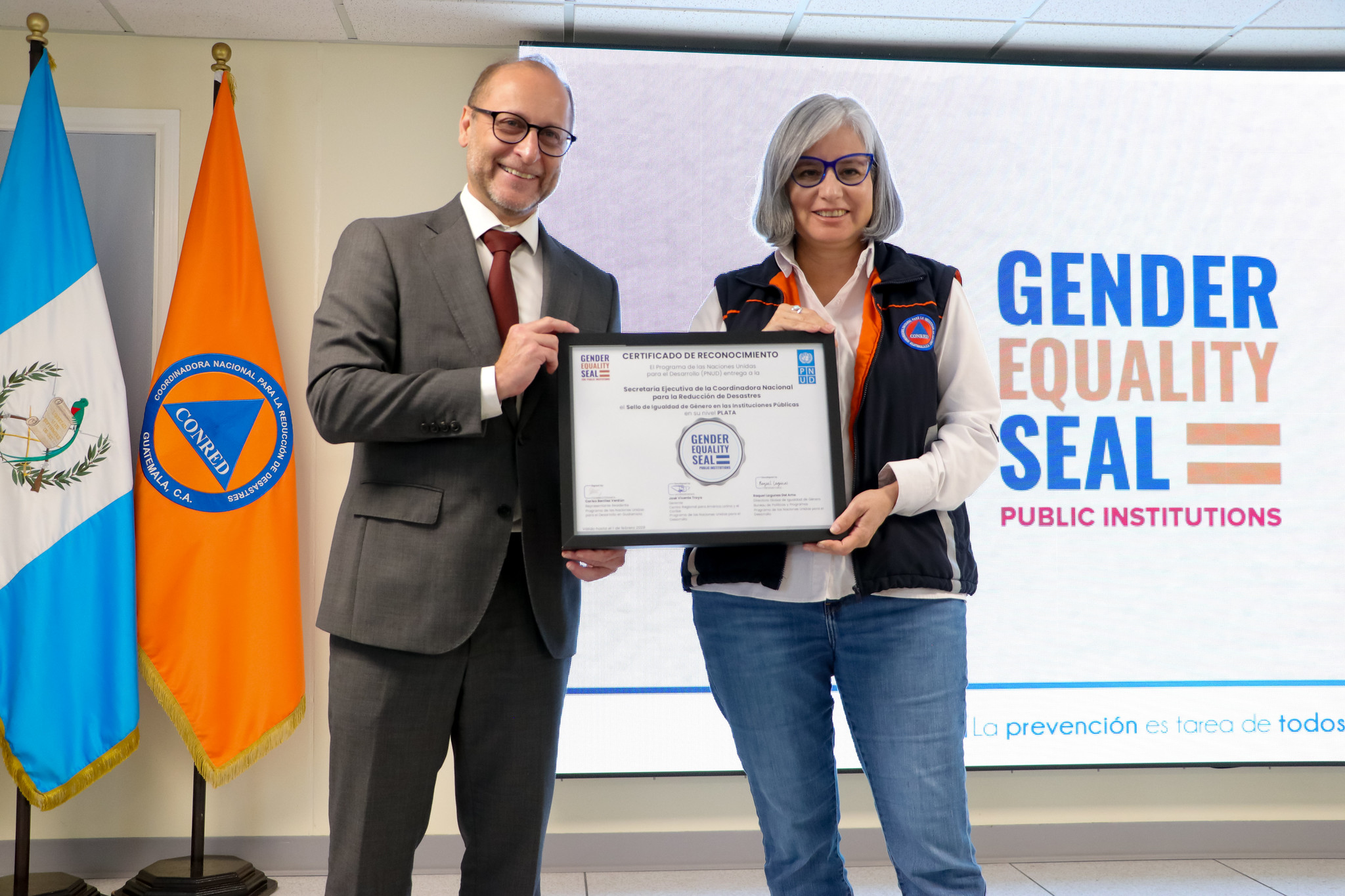 Two smiling officials hold a framed certificate on stage in front of a Gender Equality Seal banner.