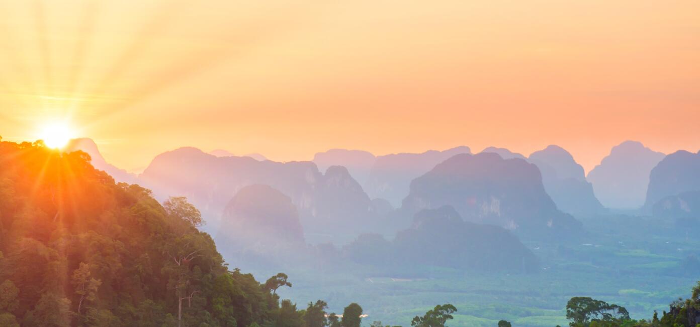 Sunrise over rolling hills toward distant blue mountains with a warm orange sky.