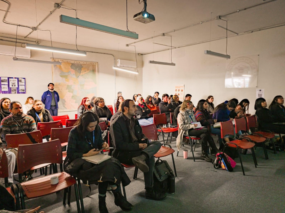 Photograph of a crowded lecture hall with red chairs and people seated, listening to a speaker.