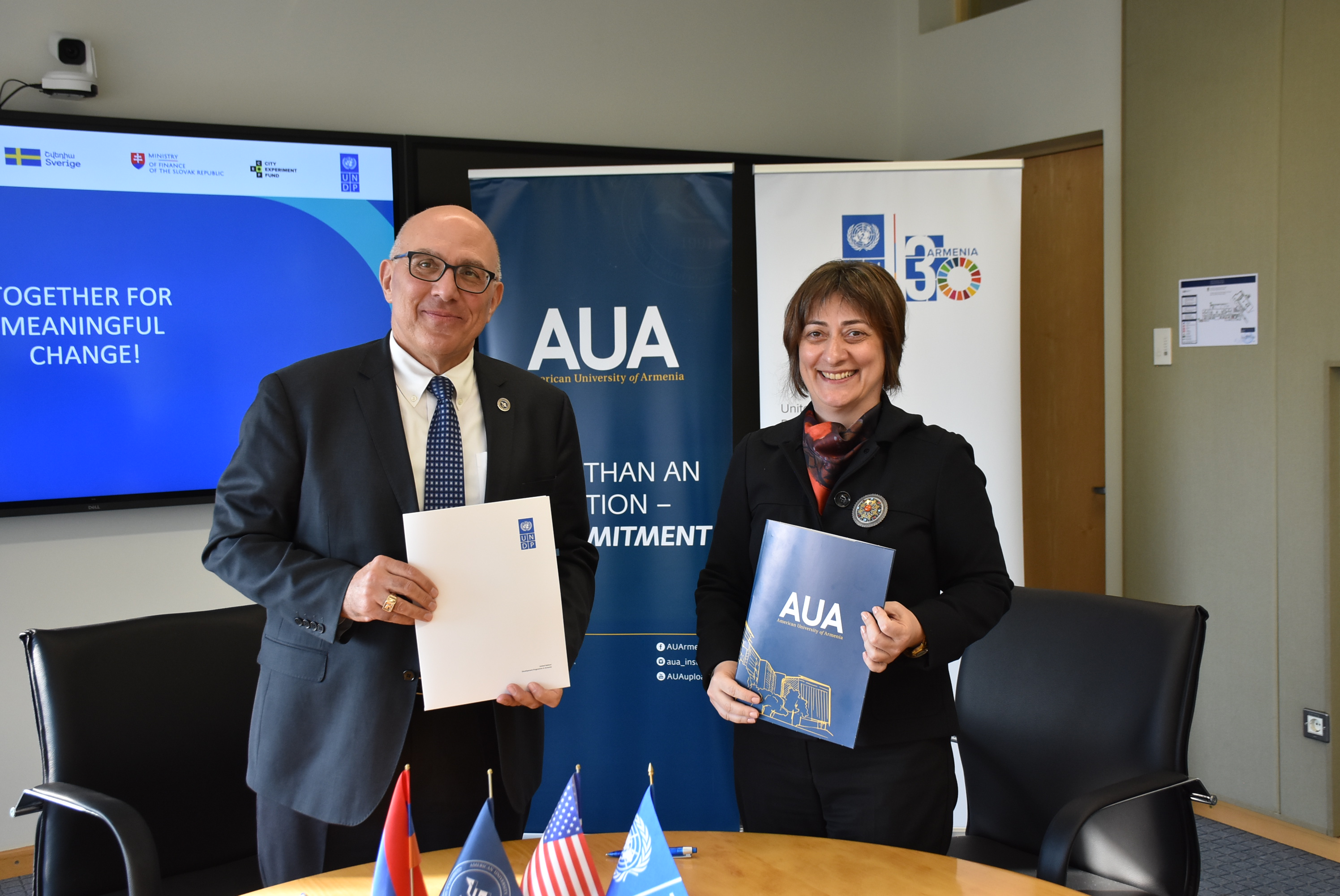 Two professionals pose with documents in a conference room, banners and flags behind them.