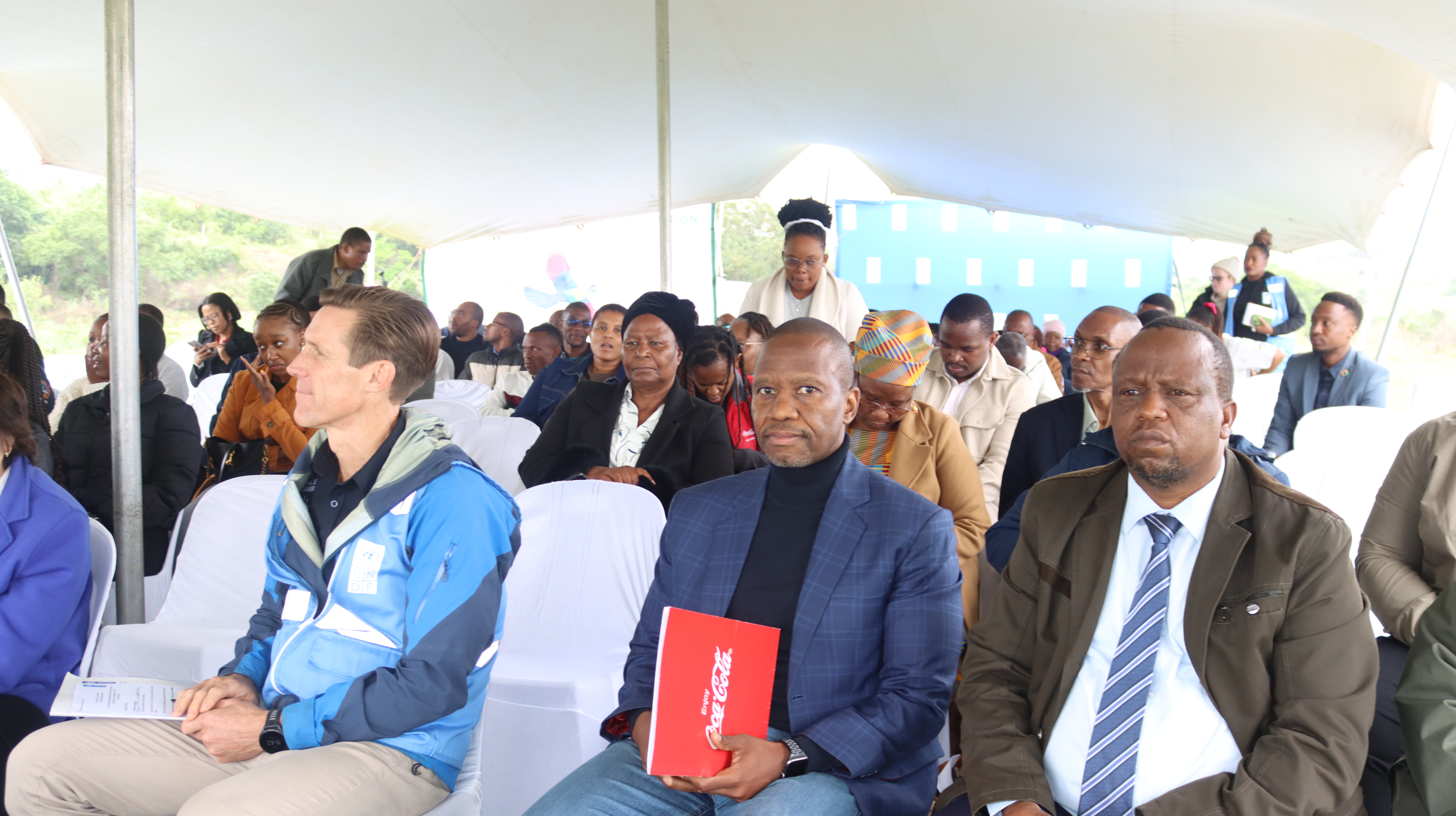 Audience under tent at an outdoor event; foreground man in suit with red folder.