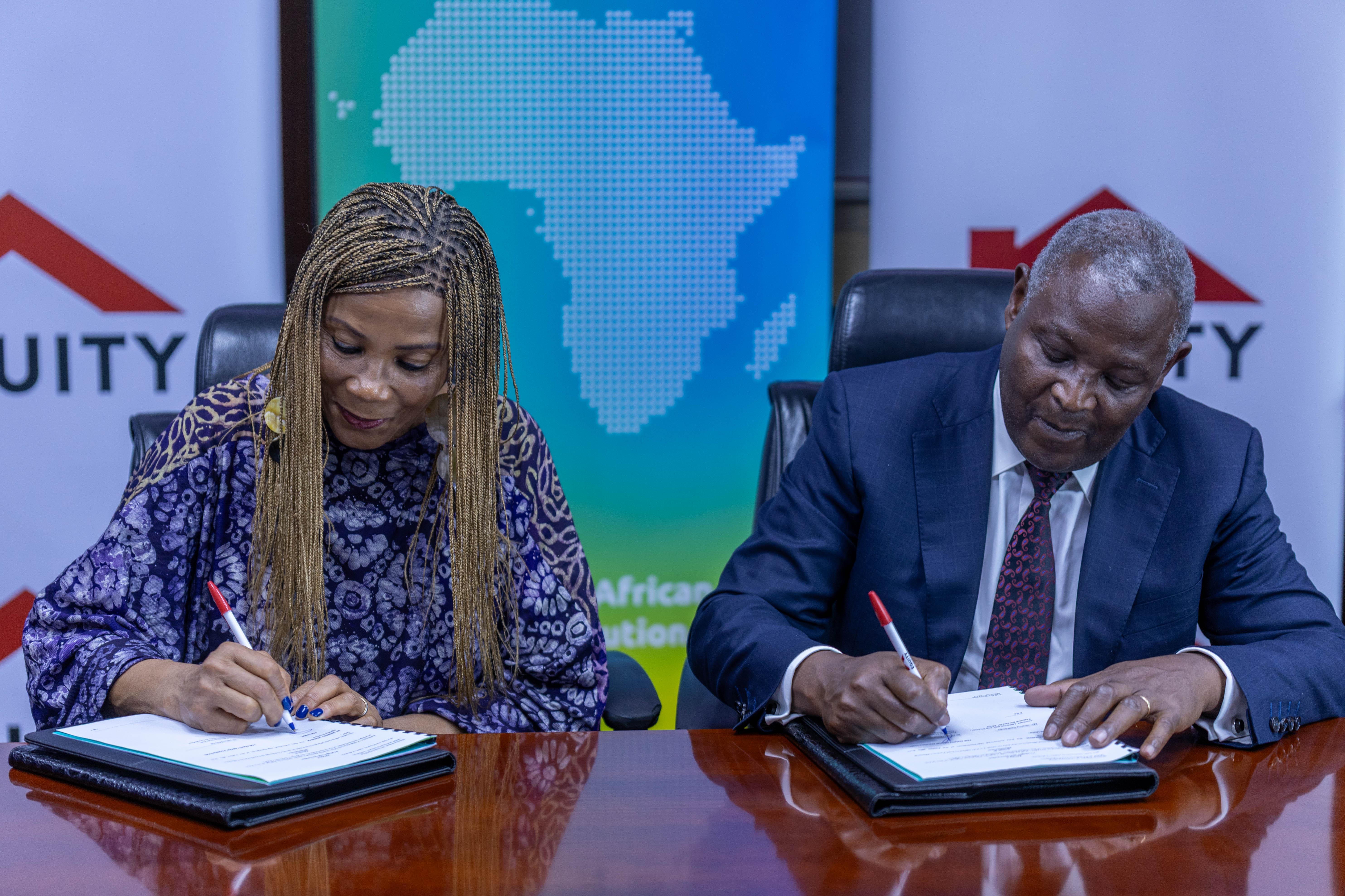 Two people sign documents at a wooden table, with blue banners in the background.