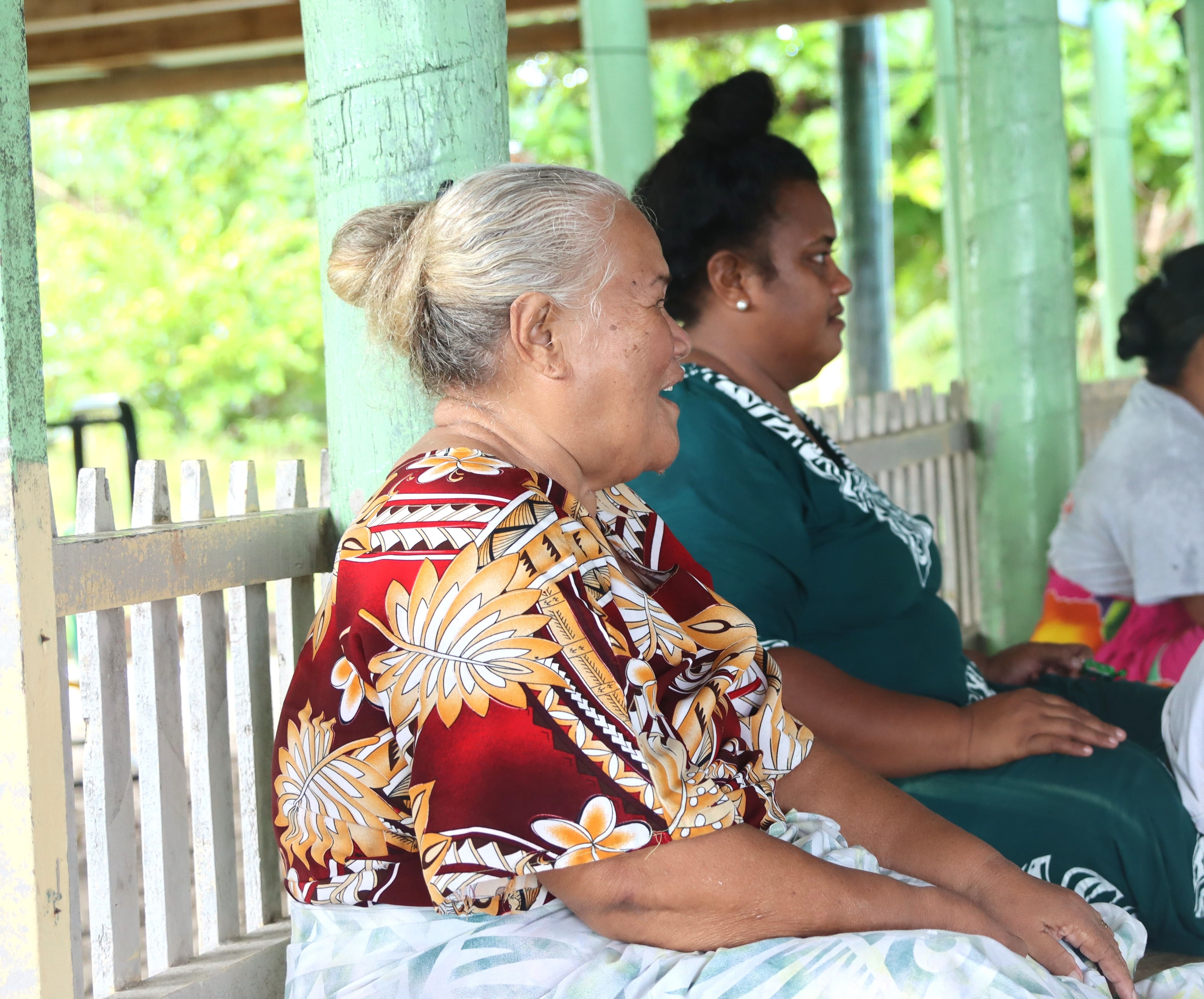 Elderly woman with gray hair in a colorful floral shirt sits on a bench under a shelter.