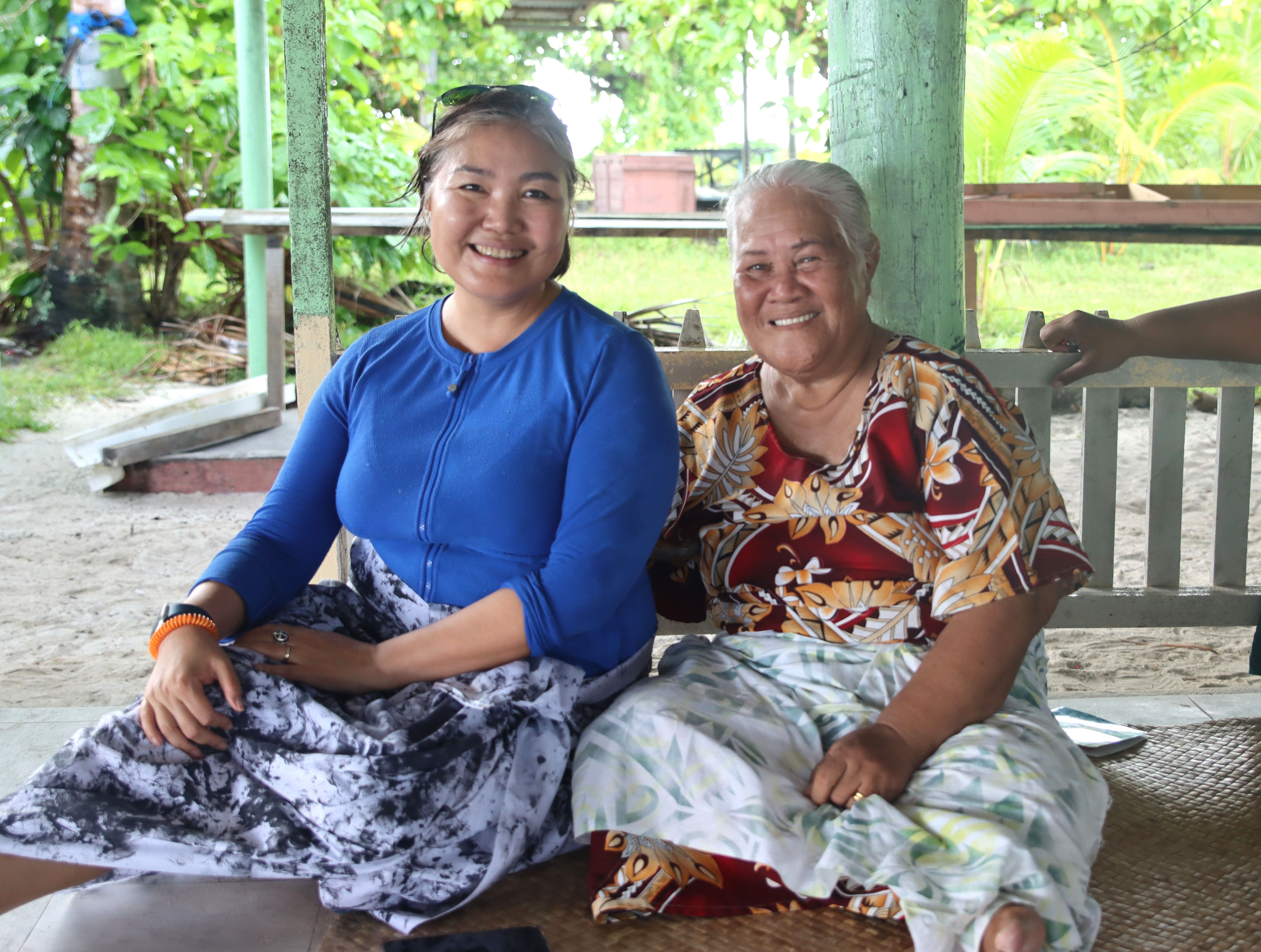 Two people on a covered porch; one in blue top with patterned skirt, the other in a colorful dress.
