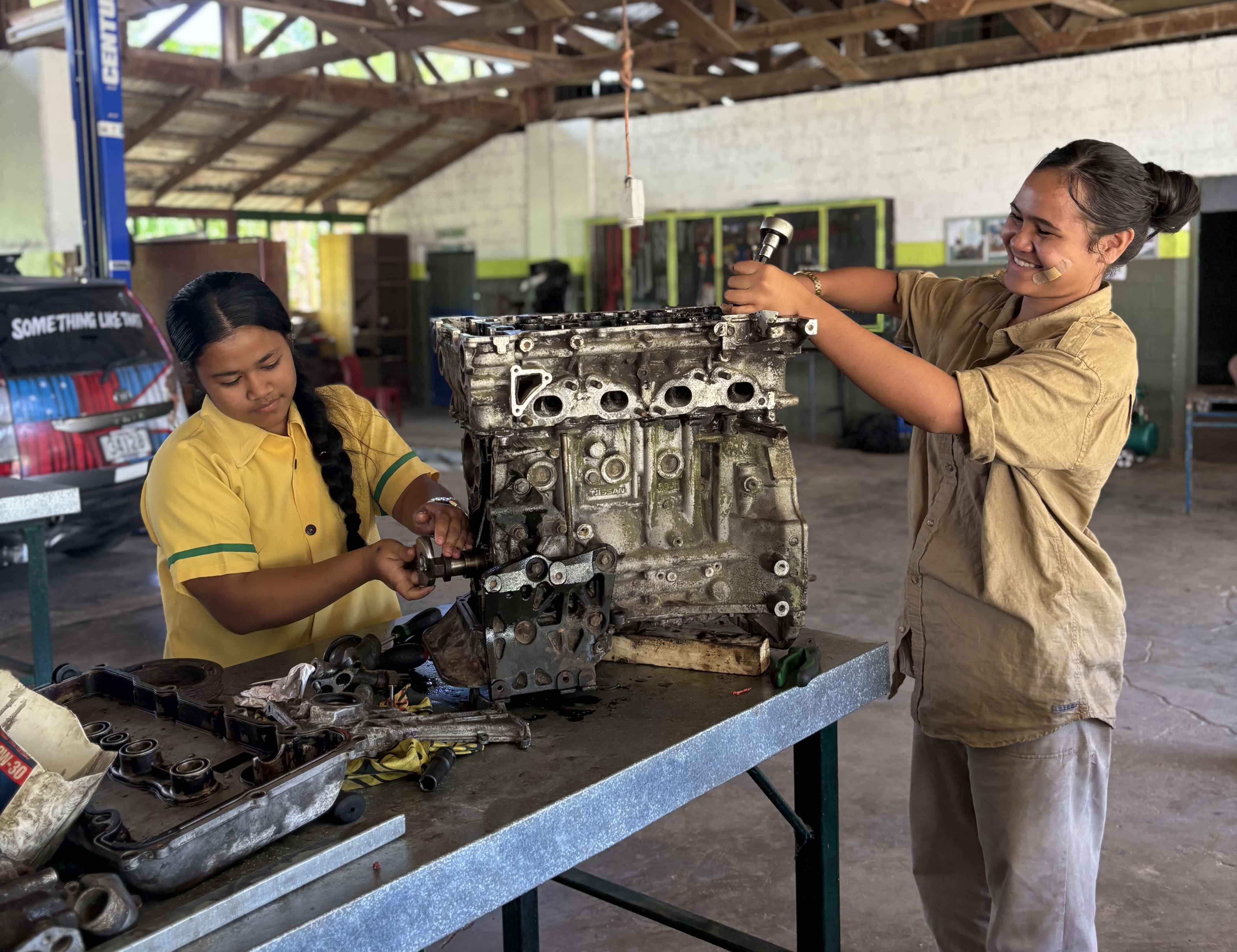 Two technicians work on a car engine on a workbench in a workshop.