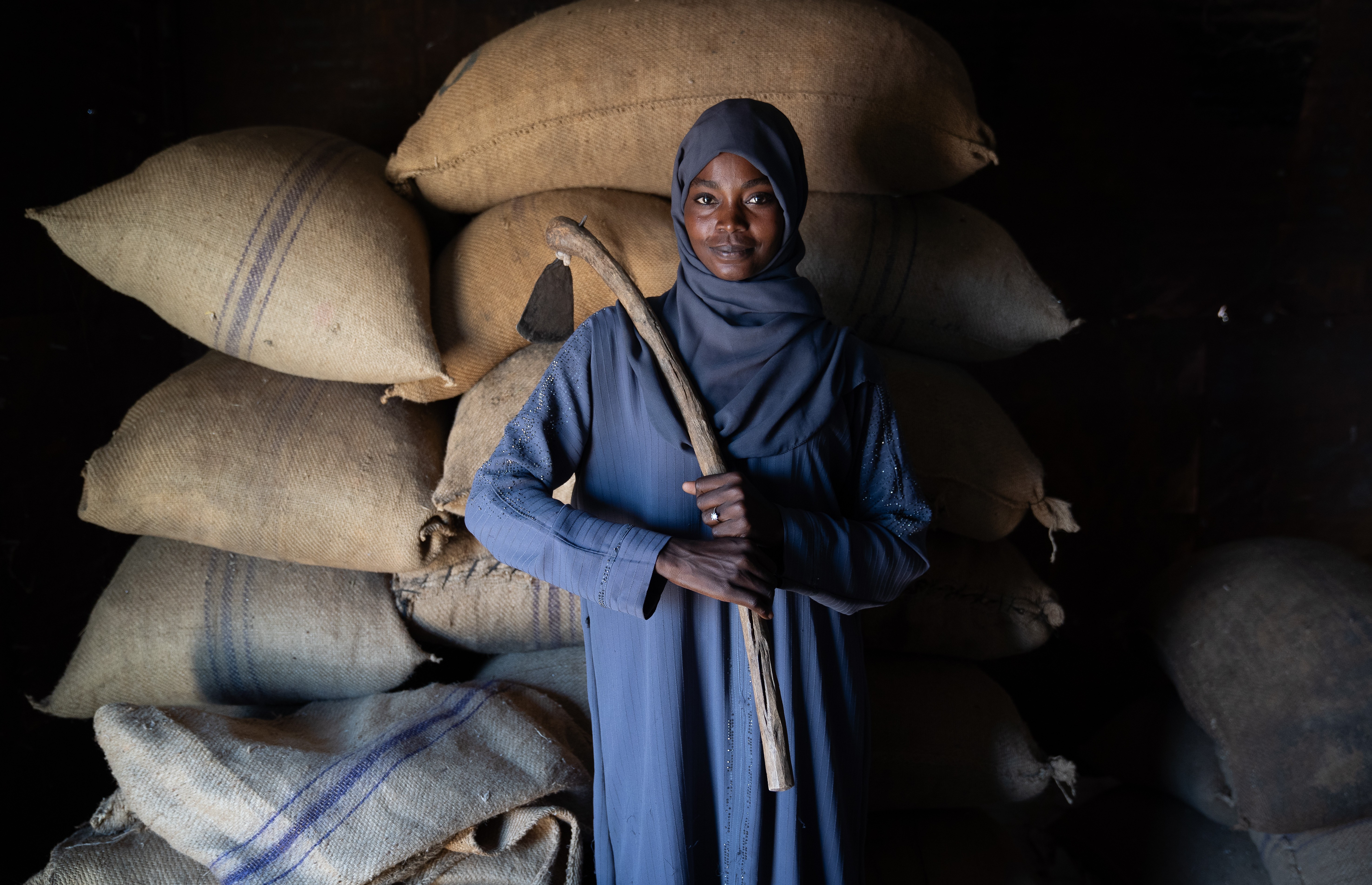 Woman in blue hijab stands beside stacked burlap sacks in a dim room.