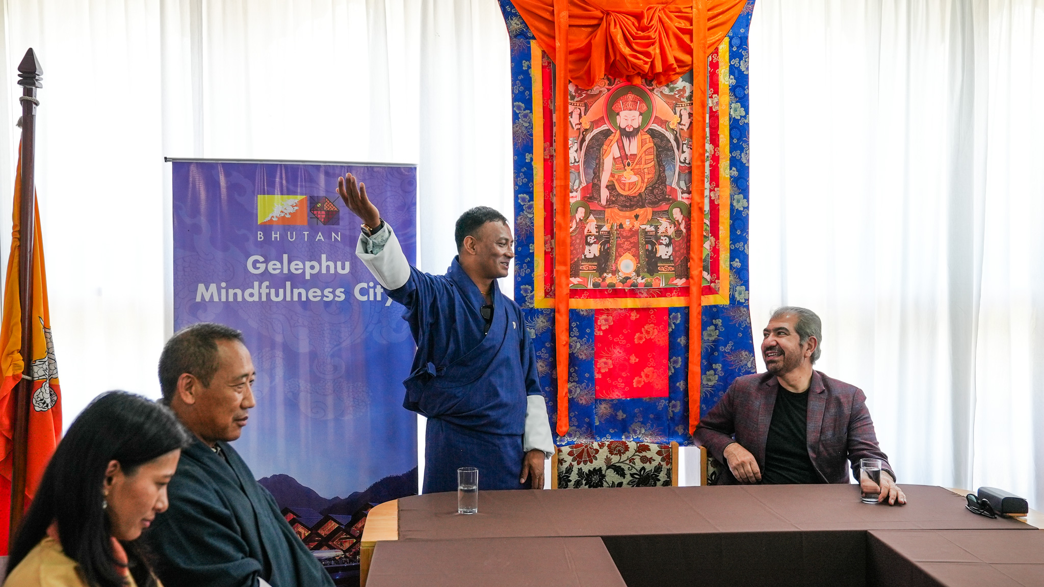 People in a conference room; a man in blue raises his arms beside a red tapestry banner.
