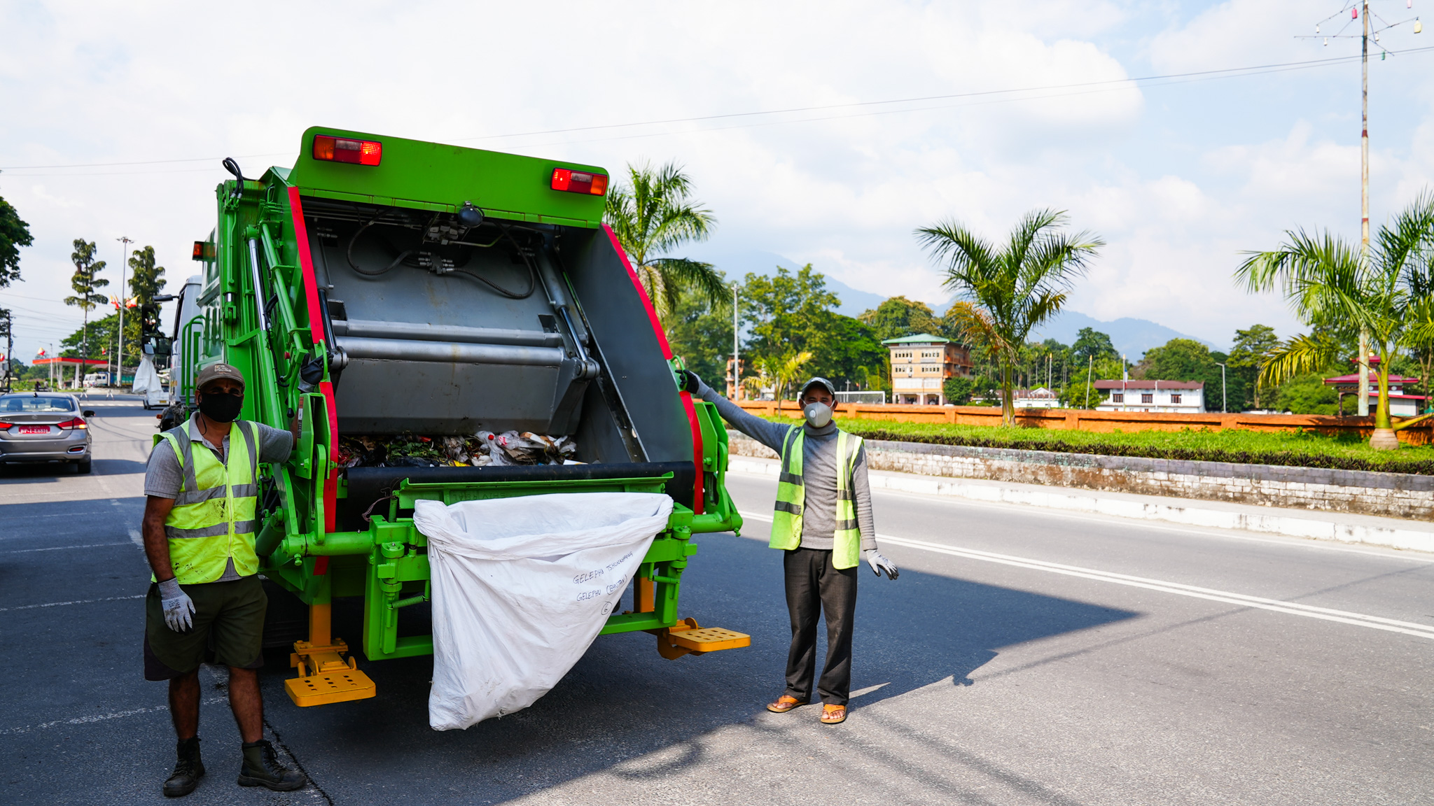 Two sanitation workers in reflective vests load a white bag into a green garbage truck on a road.