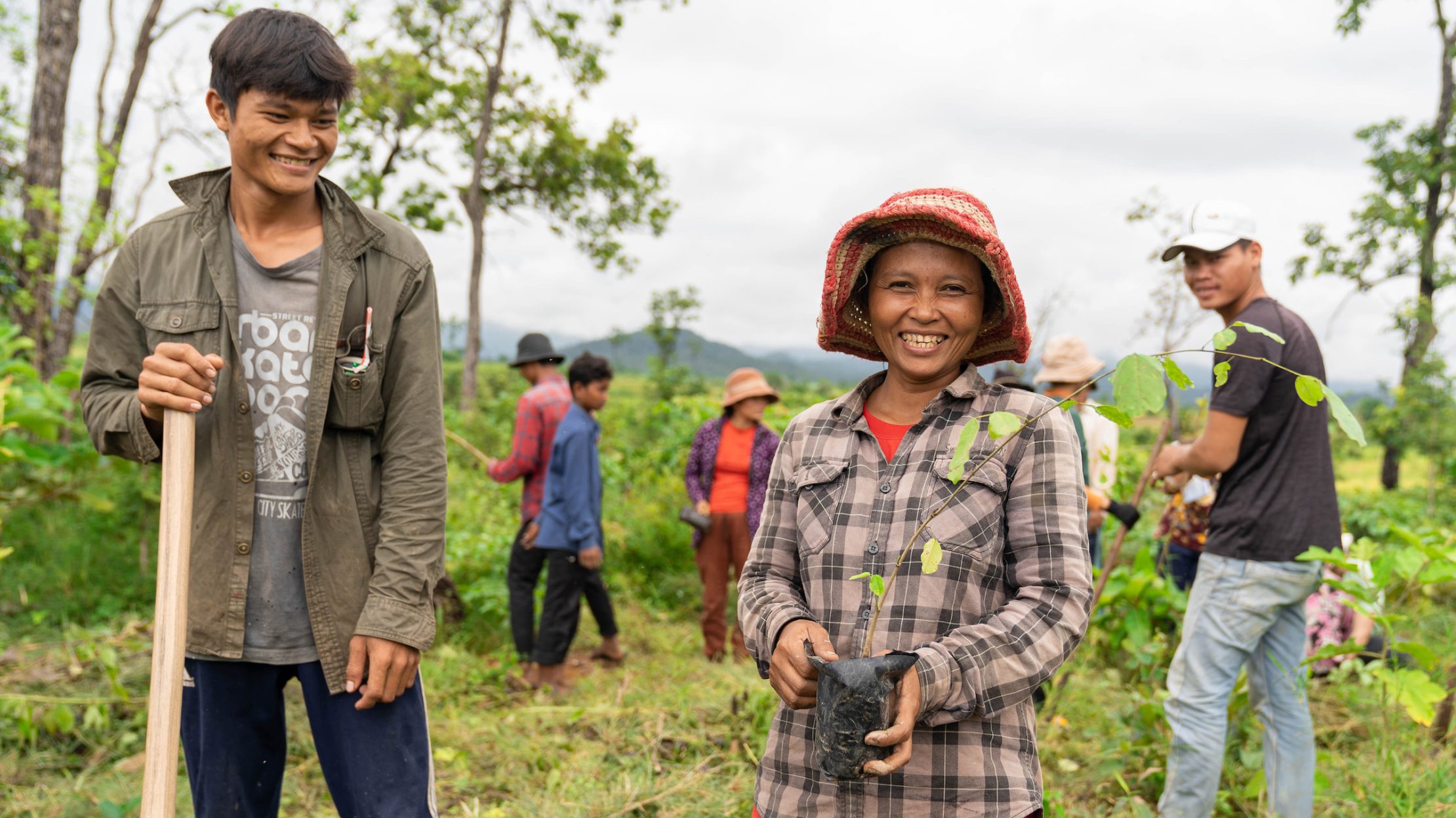 Two smiling volunteers in a field; man on left, woman in hat, others in background.