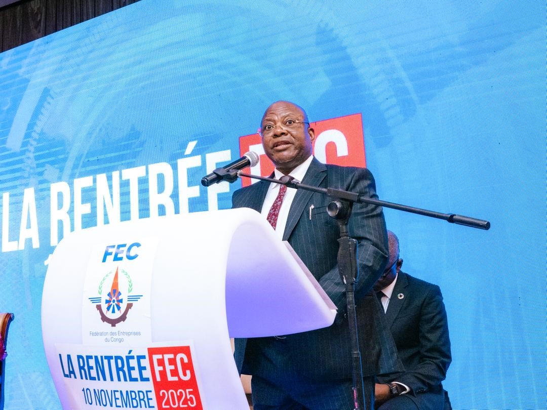 Photograph of a man in a suit speaking at a podium before a blue banner.