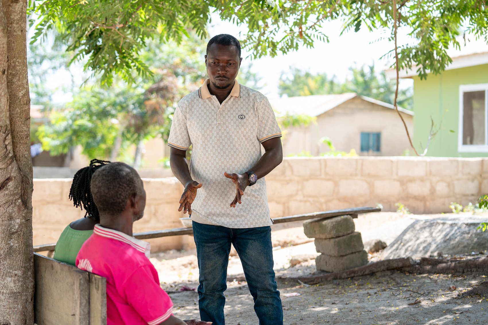 Man talks with a girl in a sunny yard; pink top, brick wall and house in background.