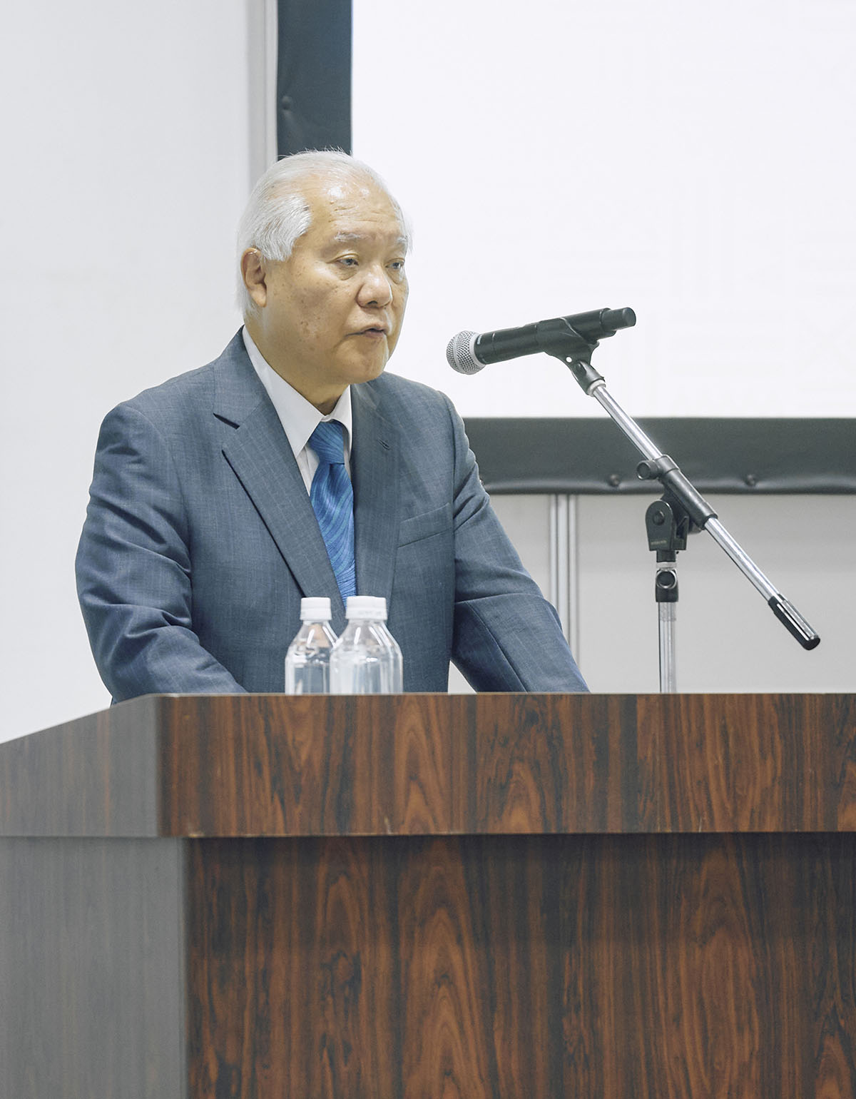 Photograph of a man in a blue suit at a wooden podium with a microphone.