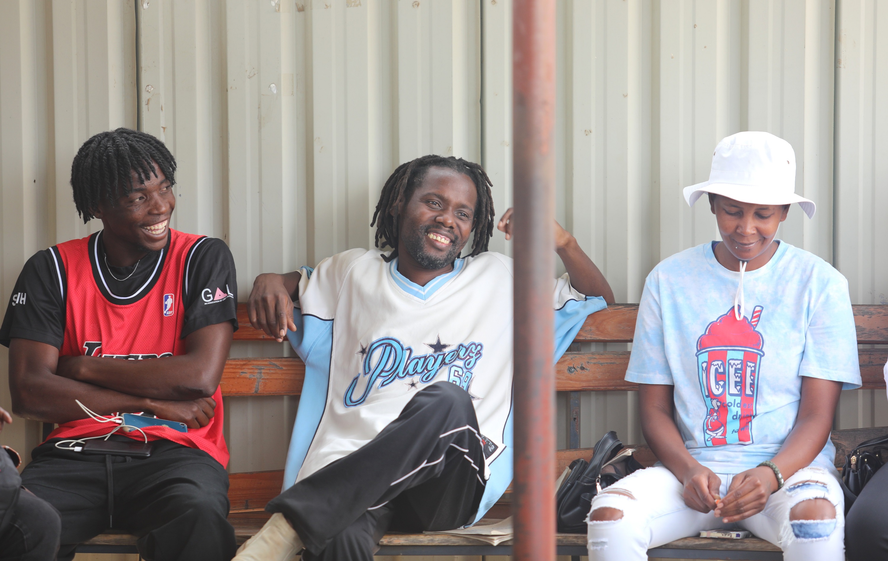 Three youths sit on a bench by a corrugated metal wall; left in red jersey, right in blue shirt.