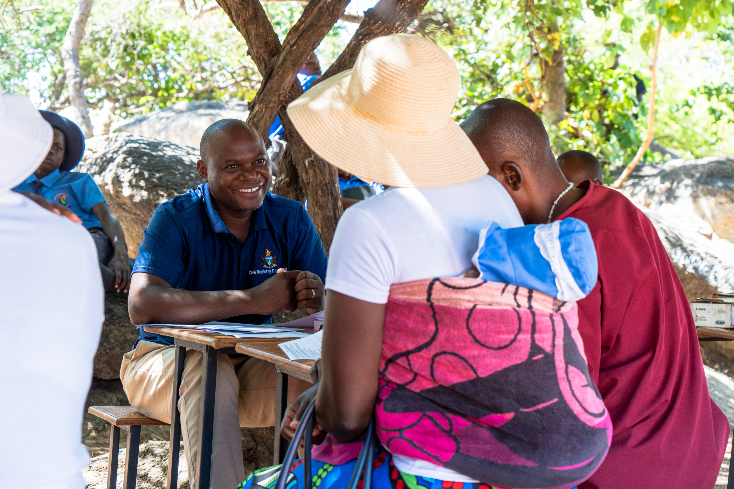 People gathered around a table outdoors under trees; man in blue shirt writes at table.