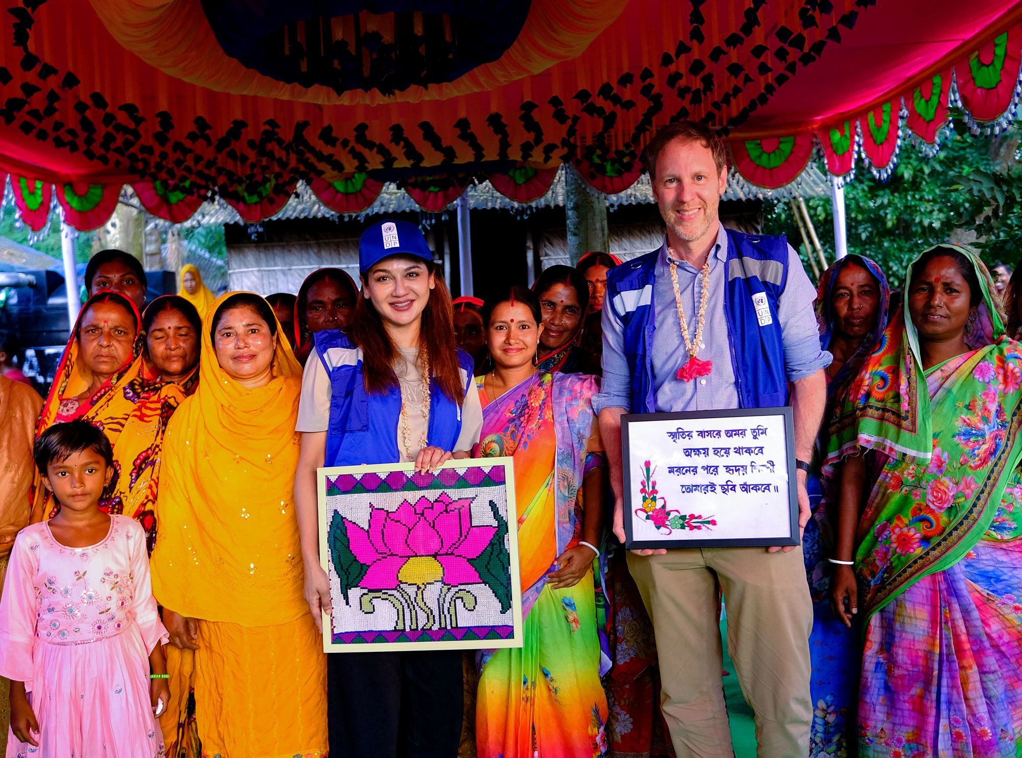 Group of people in bright clothes under a festive canopy; two adults hold framed art and a plaque.