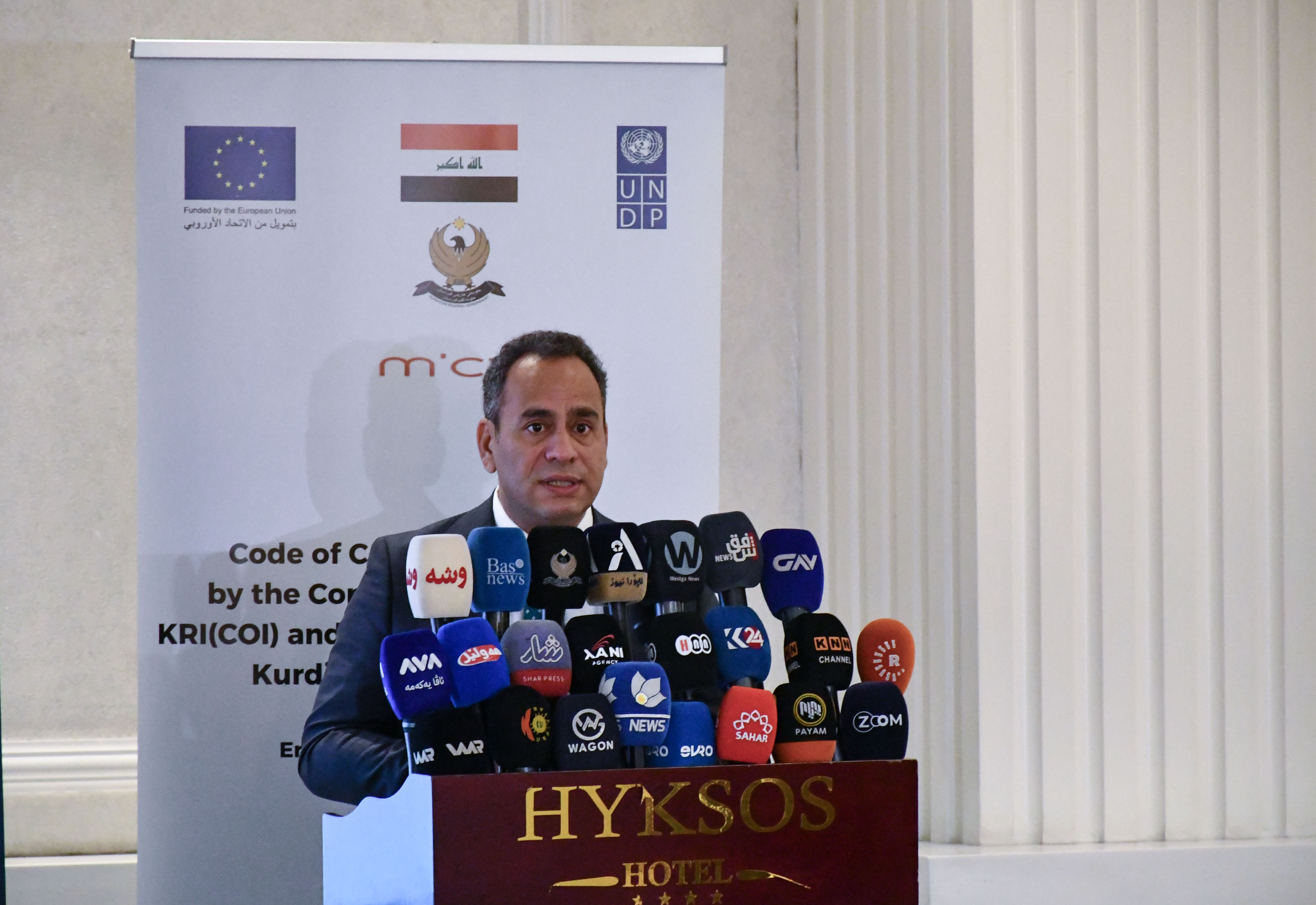 Person at a podium with many microphones; backdrop displays EU flag and organizational logos.