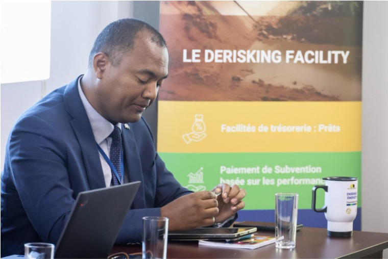 Man in blue suit at a desk with a laptop; banner behind reads Le Derisking Facility.