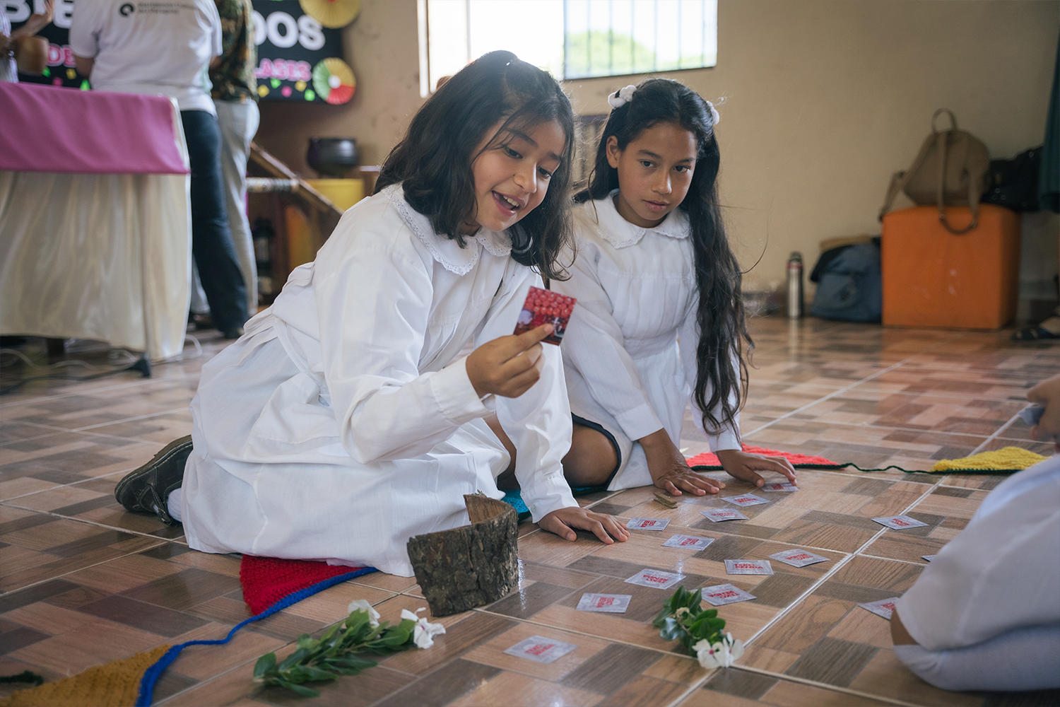 Two girls in white lab coats kneel on a tiled classroom floor, playing a memory card game with leaves and pinecones.