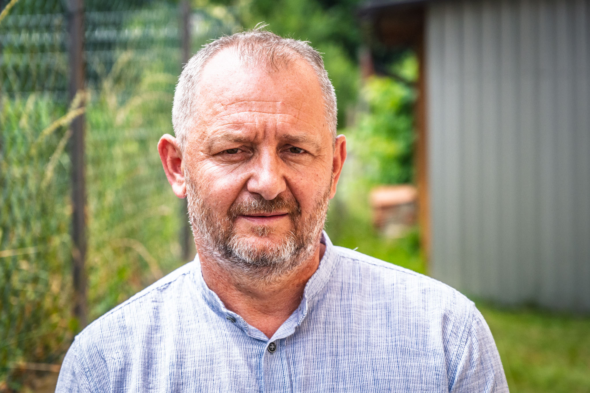 Man with gray hair wearing a light blue shirt outdoors, greenery and a shed behind.