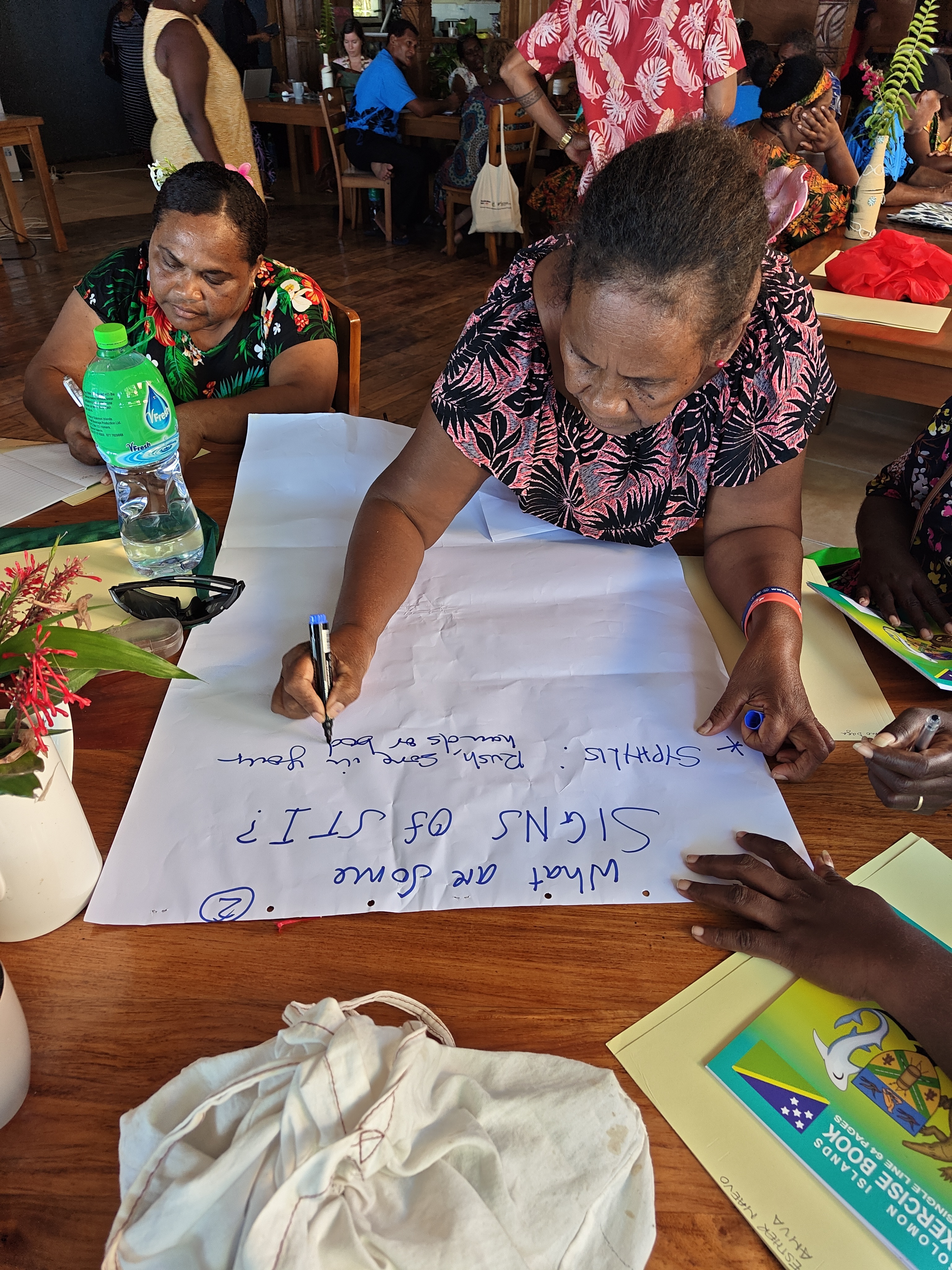 Girl in floral top writes on a large poster with a marker at a classroom table; others work nearby.