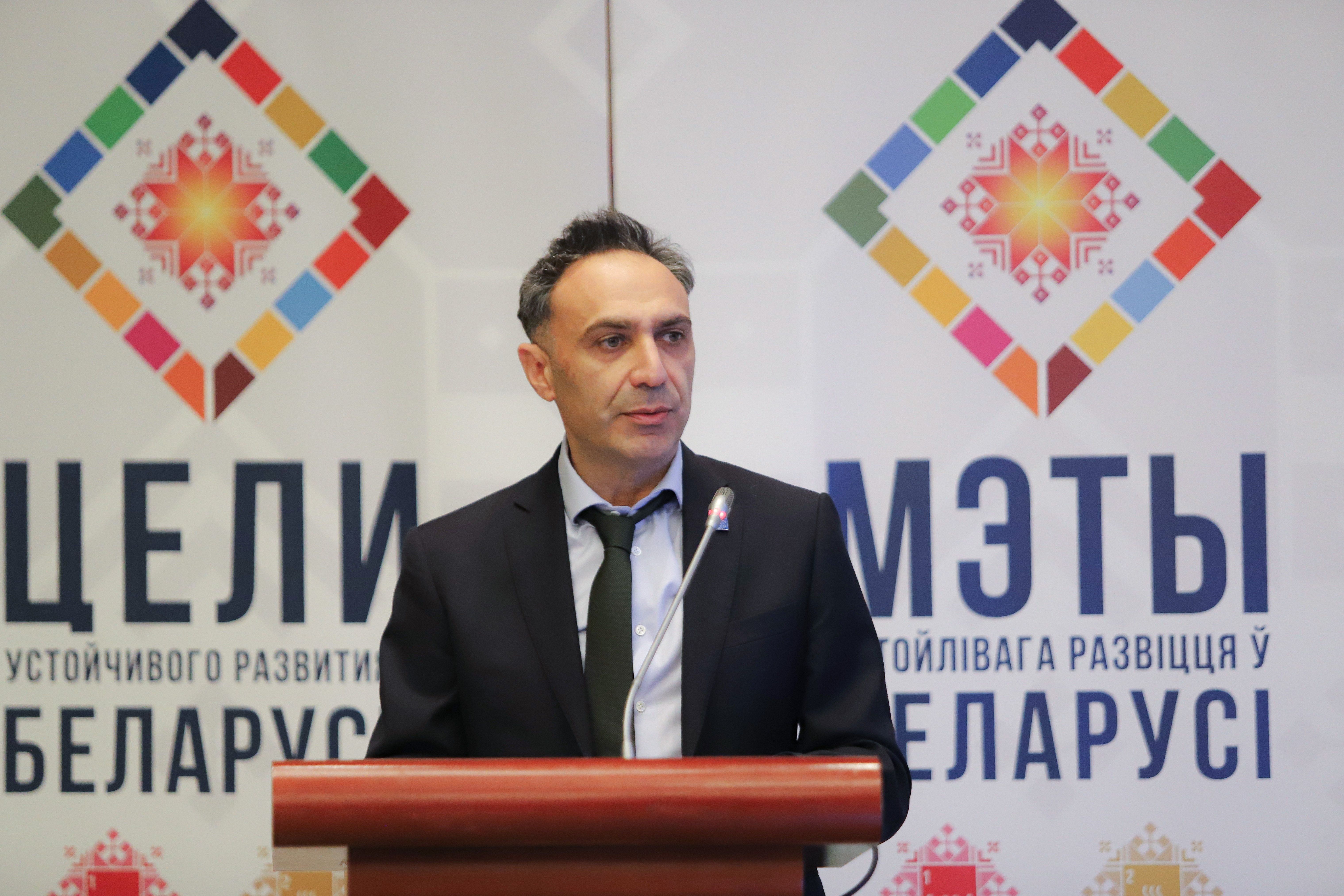 Photograph of a man at a podium with colorful diamond logos on banners featuring Belarusian text.