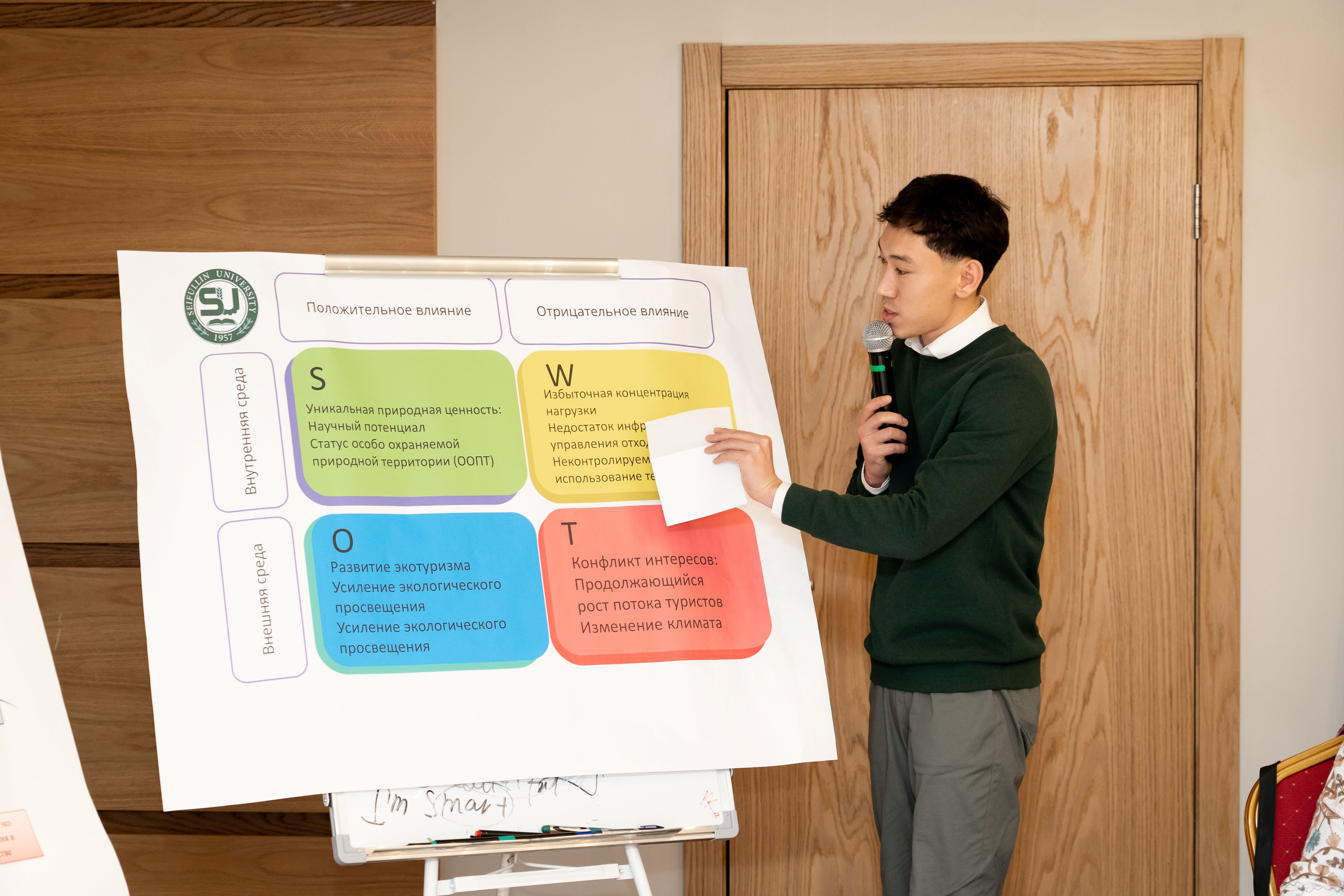 Boy in a green sweater speaks into a microphone beside a large multi-colored poster on a stand in a classroom.