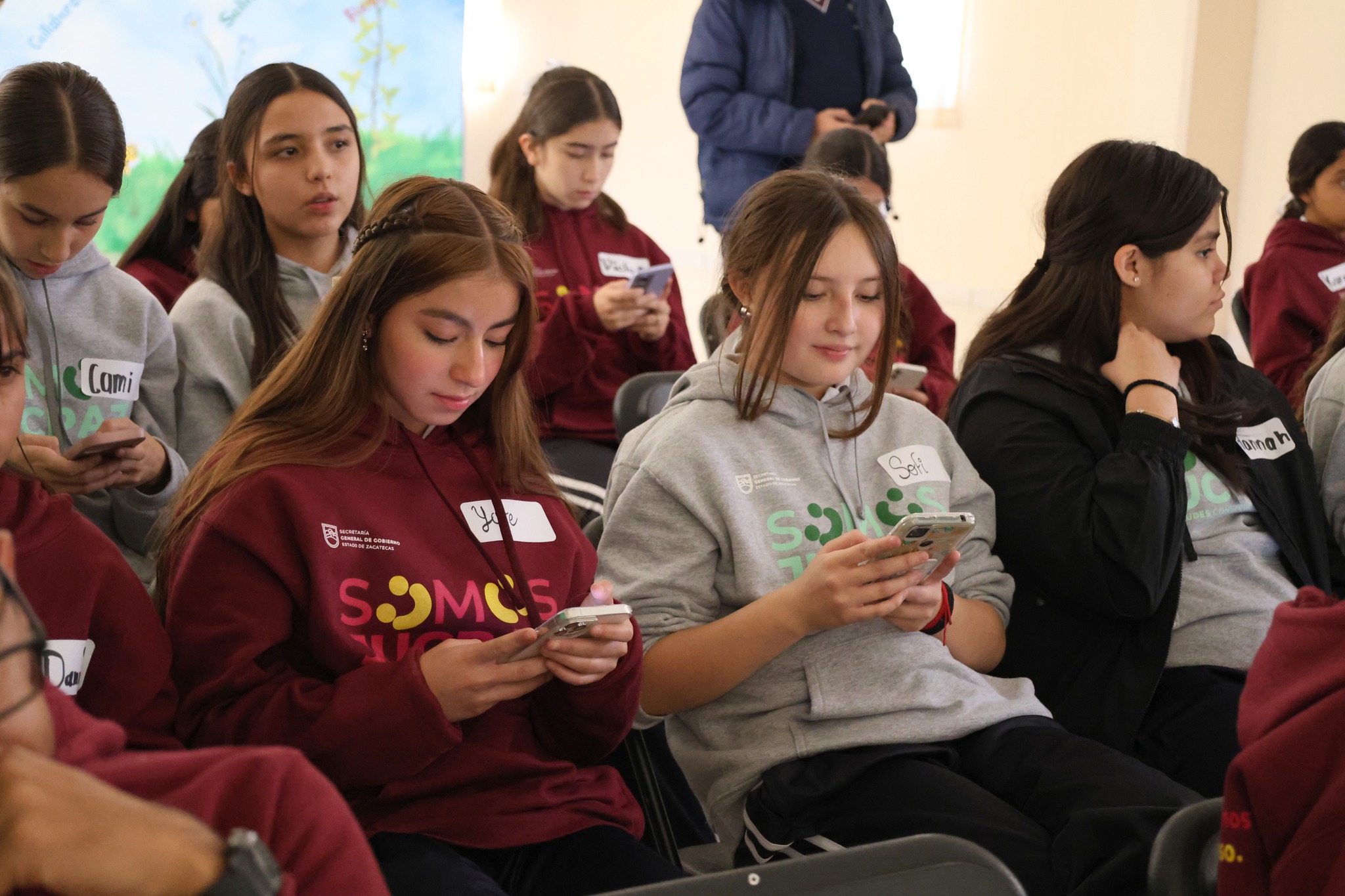 Group of students seated indoors, some looking at phones; maroon and gray shirts.