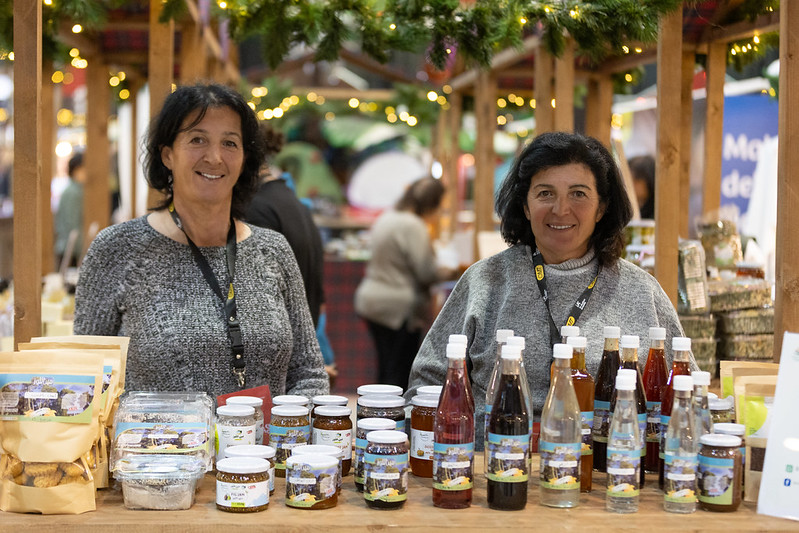Two sellers behind a wooden market stall displaying jars of preserves and bottles.
