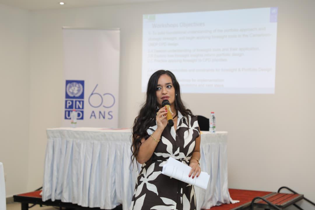 Photograph of a female presenter with microphone in conference room, slide projected behind.